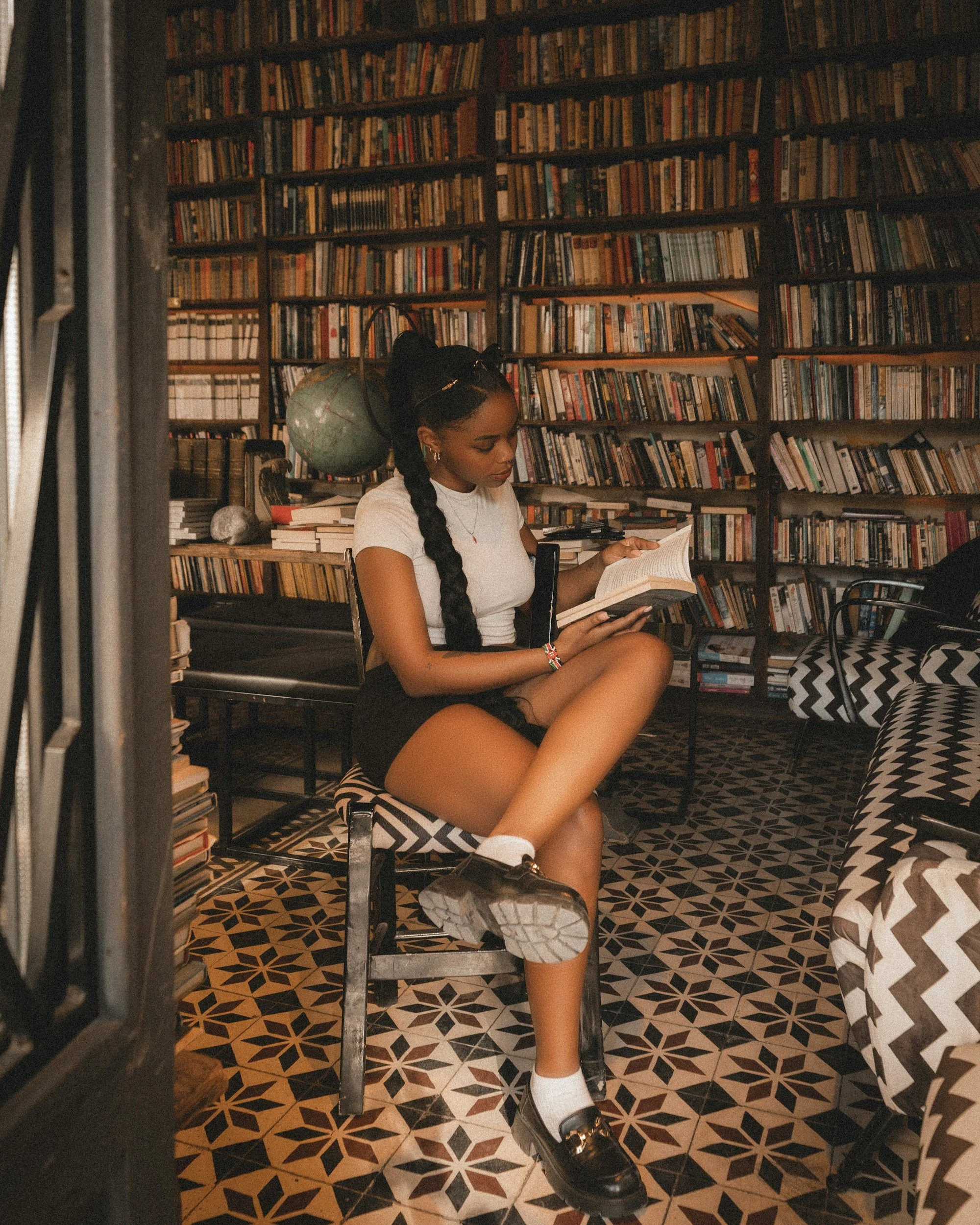 A young woman with long braided hair and hoop earrings is sitting in a cozy library, reading a book. She is wearing a white t-shirt, striped shorts, and chunky black shoes with white socks. Behind her are bookshelves filled with books, and she has a globe on her shoulder. The floor has black and beige geometric patterned tiles, and there is a black and white zigzag cushion nearby.