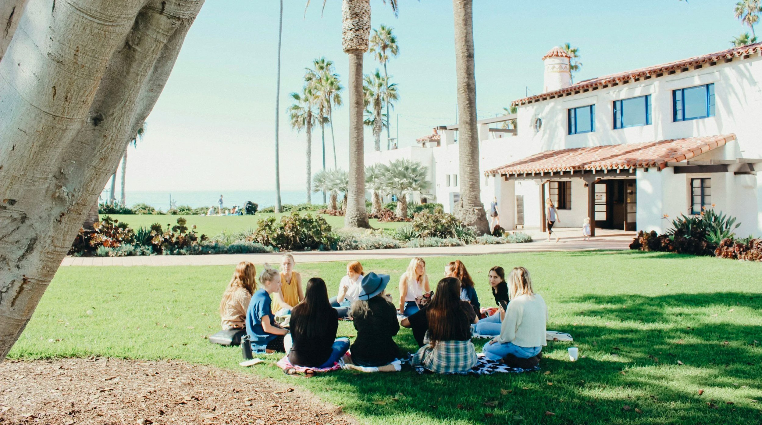A group of people sitting on a blanket on green grass in a park, with palm trees, a white building with red tile roof, and a clear sky in the background.