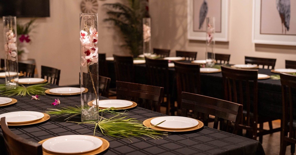 Dinner table set with white plates with gold rims, and decorative tall glass vases with orchids and greenery, in a warmly lit room.
