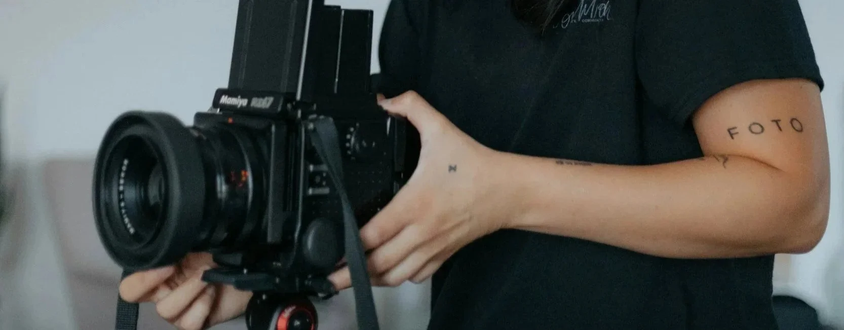Person holding a large professional camera with a black body and lens, wearing a black shirt, tattooed arms with tattoos reading 'FOTO' and a small star, in an indoor setting.