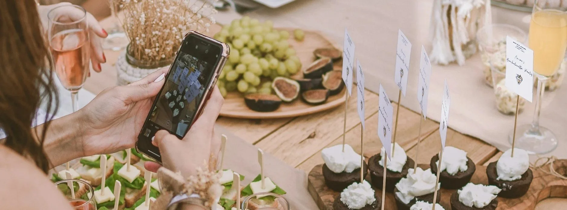 Person taking a photo at a dessert table with various treats, grapes, figs, and small flags with writing