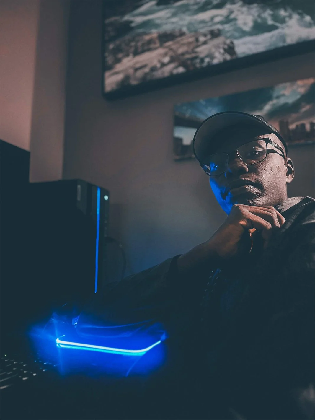 A young man wearing glasses and a cap sitting in a dimly lit room with a computer, blue LED lighting, and framed landscape photographs on the wall.