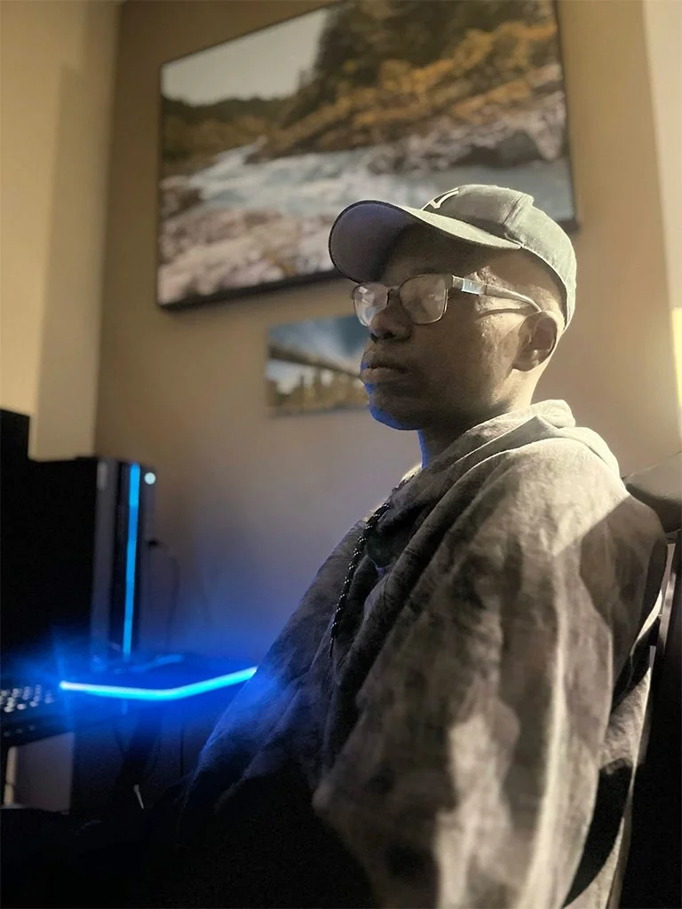 A young person wearing glasses, a green baseball cap, and a hoodie sitting in front of a computer with a blue light. There are landscape photographs on the wall behind them.