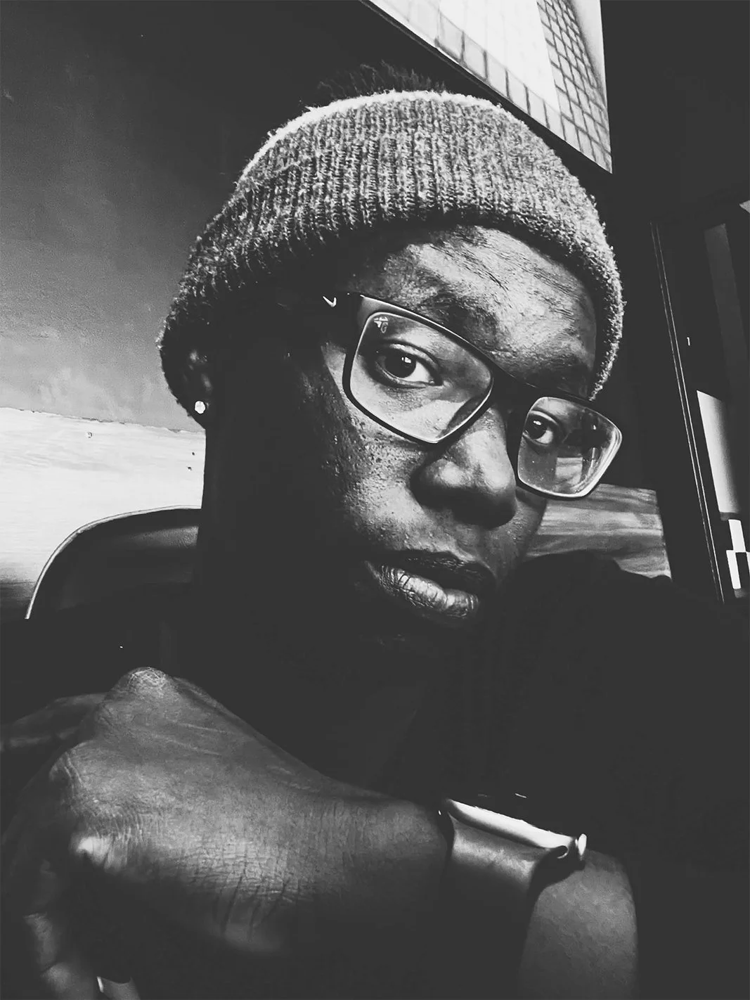 Black and white close-up photo of a woman wearing glasses and a beanie, resting her chin on her hand, with a textured wall in the background.