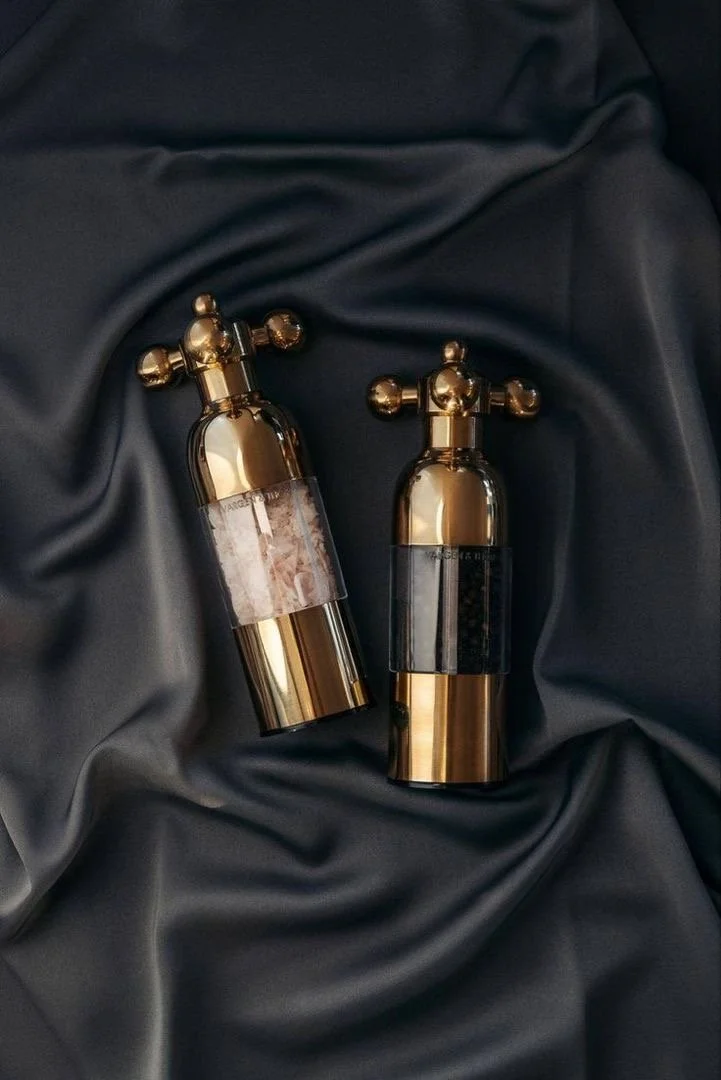 Two luxurious gold-colored spray bottles with decorative crosses on top, resting on dark, silky fabric.