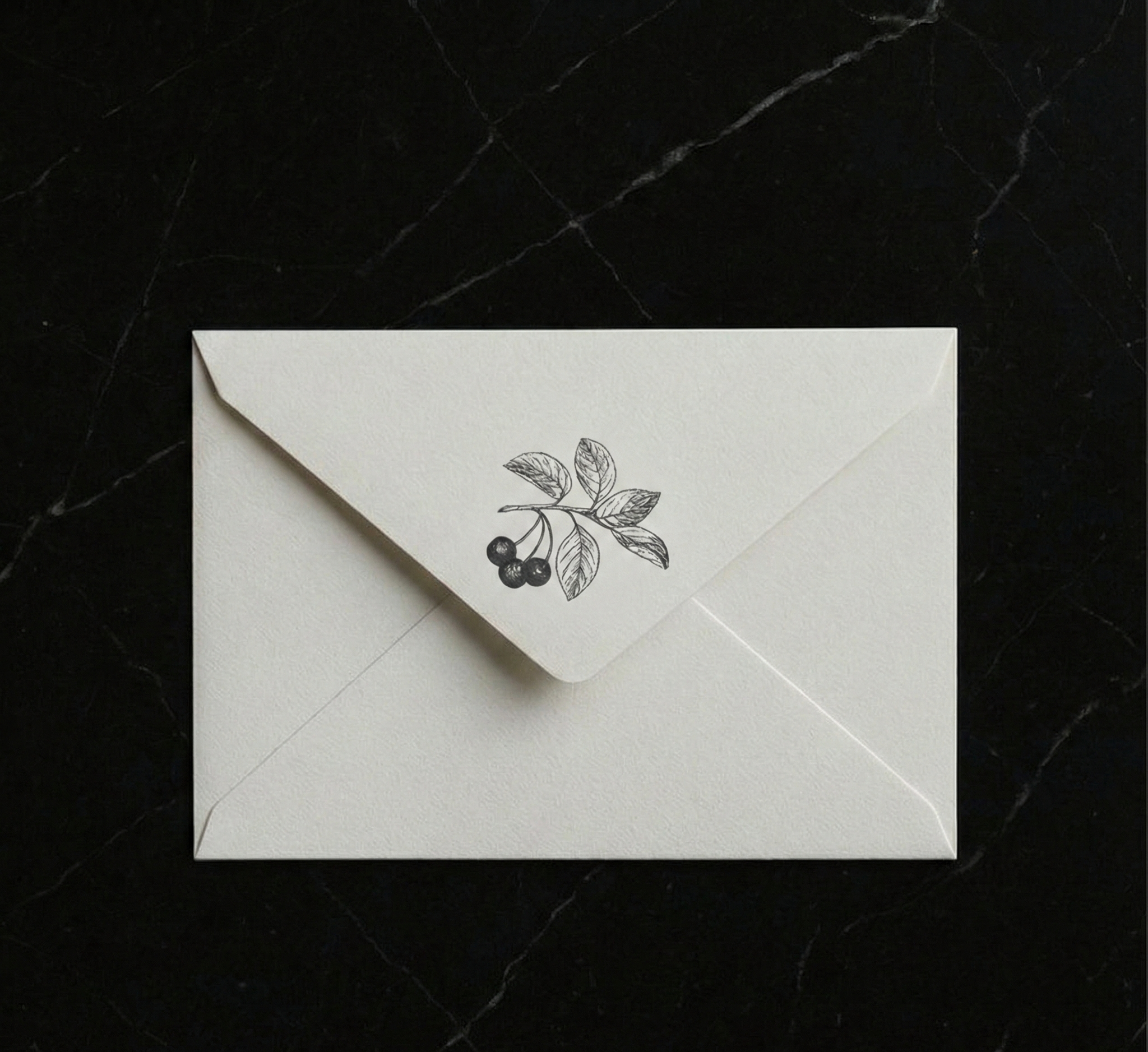 A white envelope with a black illustration of a branch with leaves and berries on the flap, placed on a black marble surface.