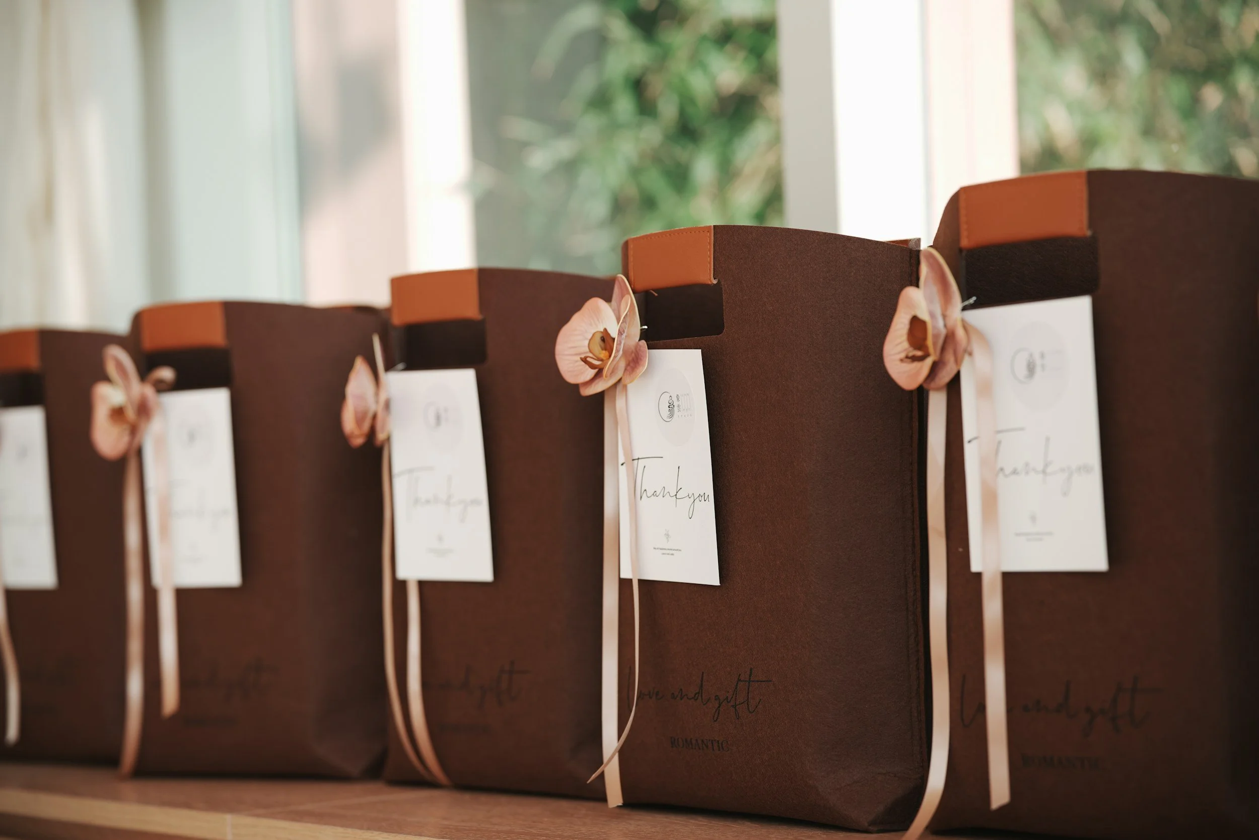 Several brown paper gift bags with pink flowers and white thank you tags lined up on a wooden surface near a window.