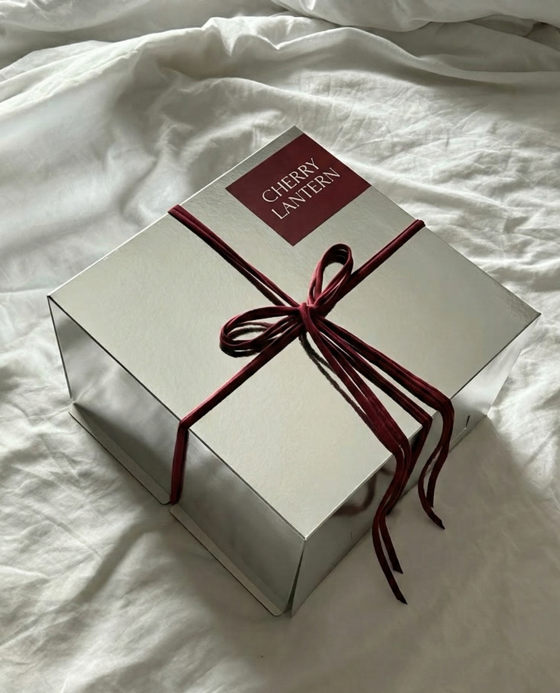 A white box labeled 'Cherry Lantern' wrapped with a burgundy ribbon tied in a bow, resting on a white fabric surface.
