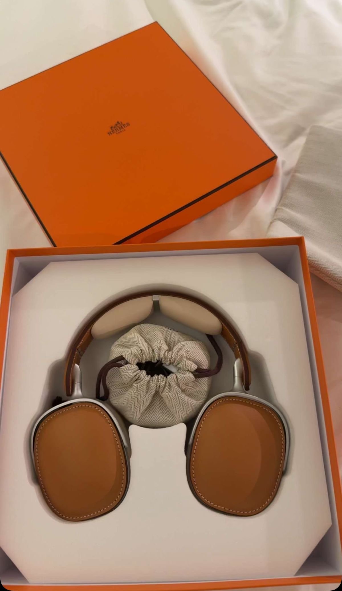 A pair of Hermes headphones with brown leather ear cups and beige headband, inside an orange Hermes box with a cloth pouch beside it.