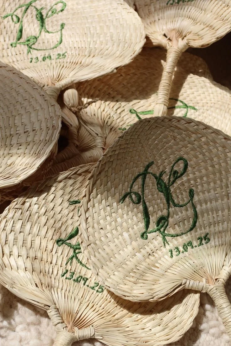 Multiple woven straw fans with green embroidered initials and date, arranged in a pile.