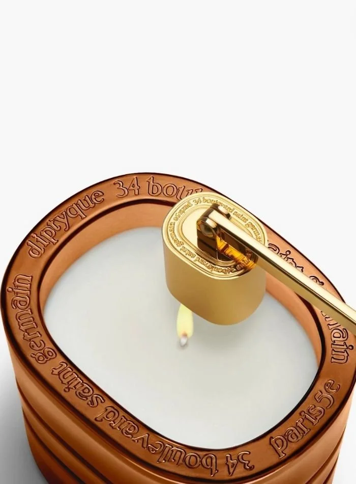 Close-up of a tea light candle inside a copper-colored holder with French text around the edge, and a gold matchstick being used to light it.