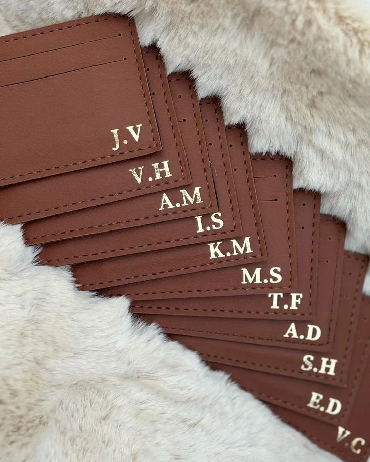 A series of brown leather cardholders, each with initials in white, arranged in a cascading pattern on a white fuzzy surface.