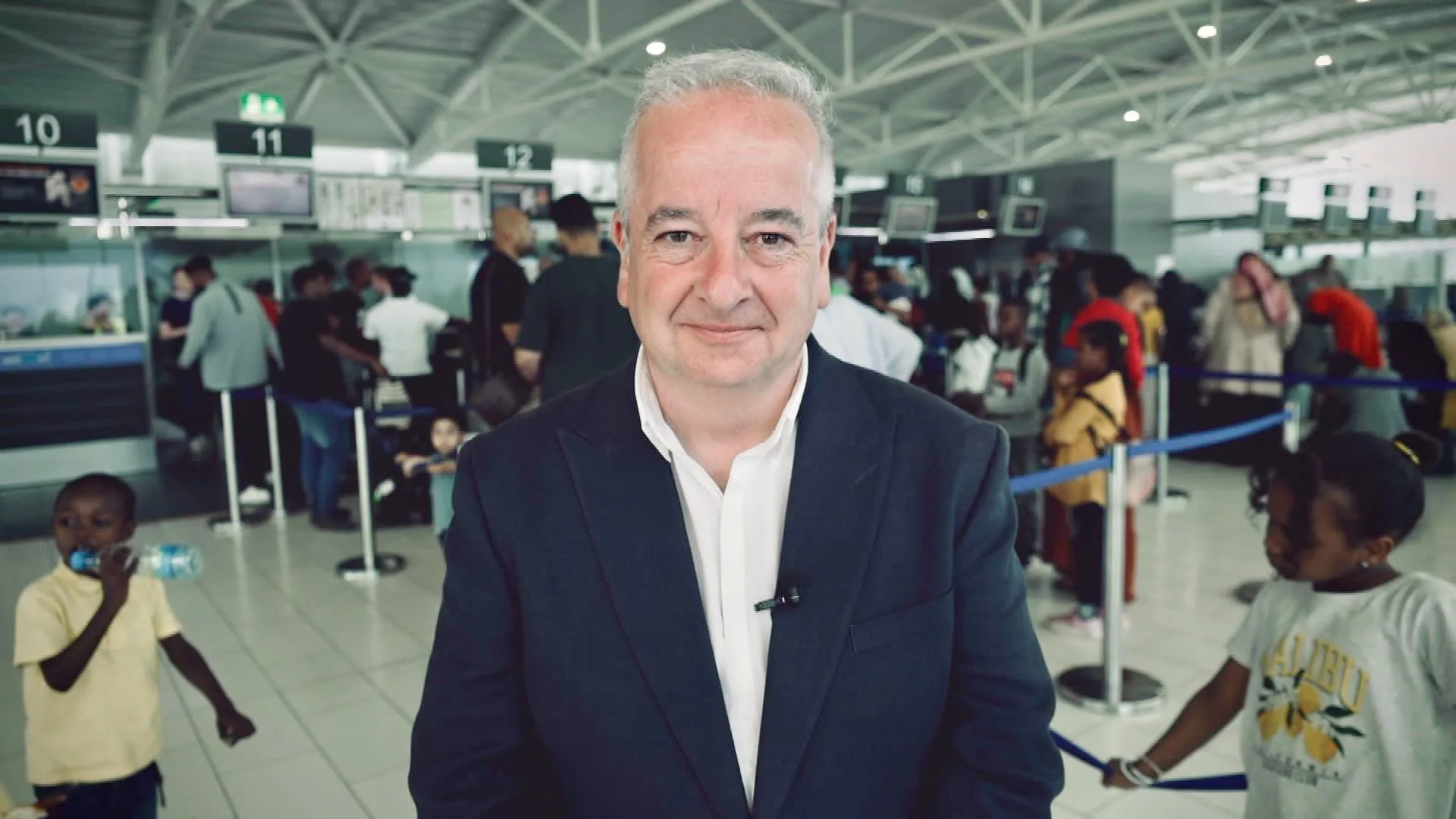 A middle-aged man in a black suit with a white shirt standing at an airport, with people in the background waiting in line and walking around.
