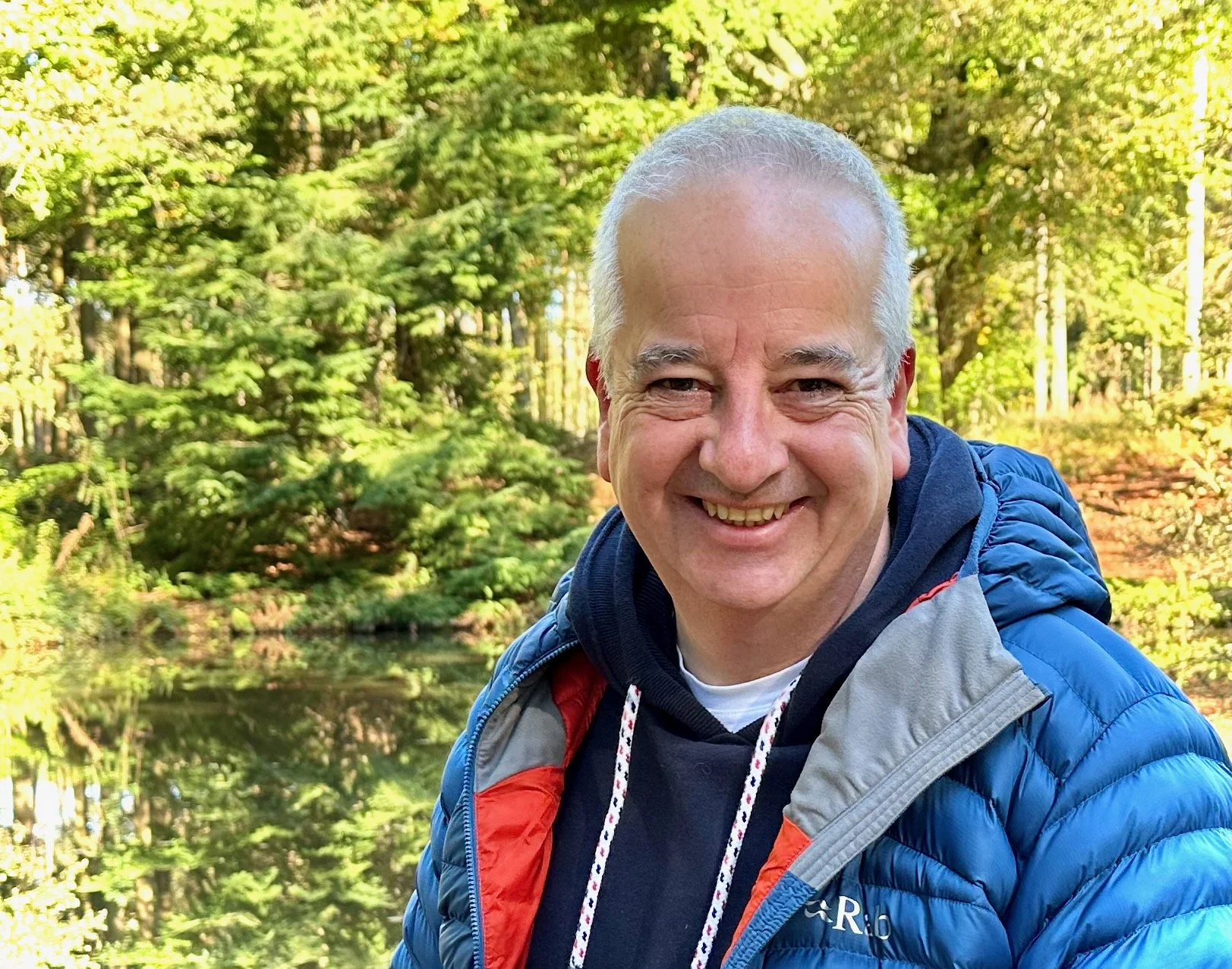 A smiling middle-aged man with short gray hair outdoors in a forested area with green trees and a pond in the background.