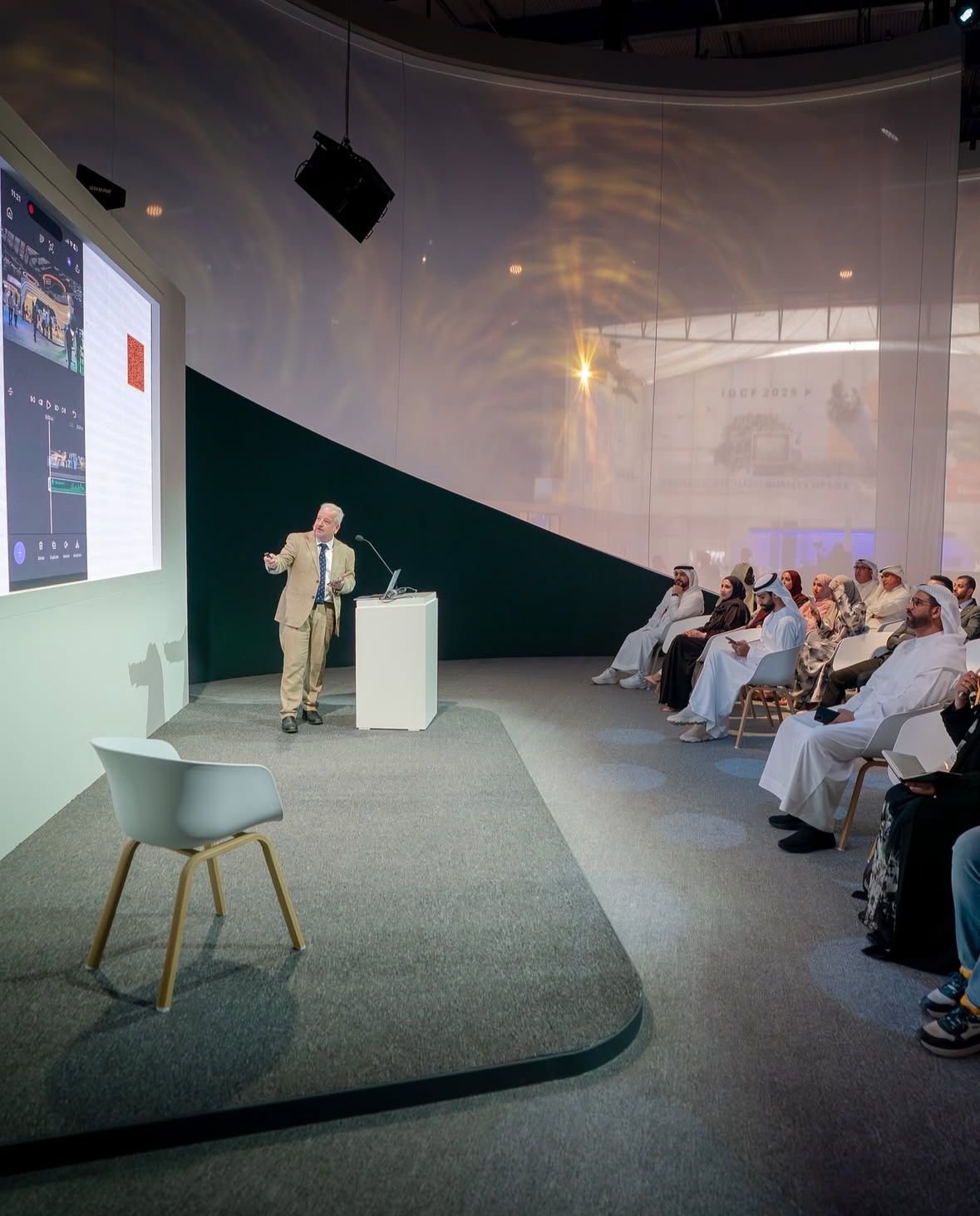 A man in a tan suit giving a presentation to an audience seated in a modern, curved auditorium. The presentation is displayed on a large screen on the left side of the image.