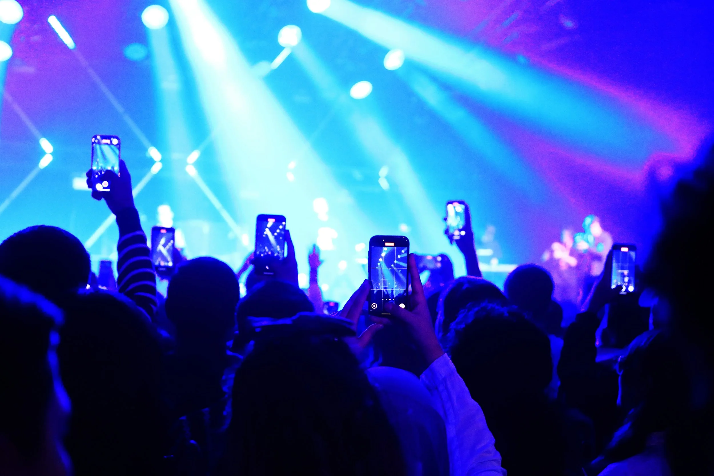 People at a concert taking photos and videos with their smartphones, illuminated by blue and purple stage lights.