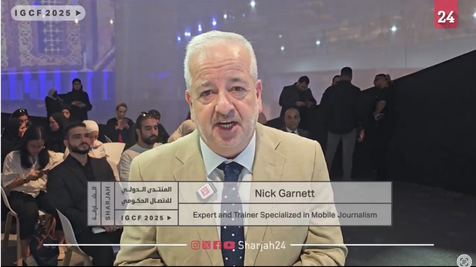 A man with gray hair and beard, wearing a beige blazer, white shirt, and navy blue tie, speaking at a conference or event. Behind him are seated people and a large screen displaying event information, with some attendees standing and talking. The event appears formal, with a diverse audience.