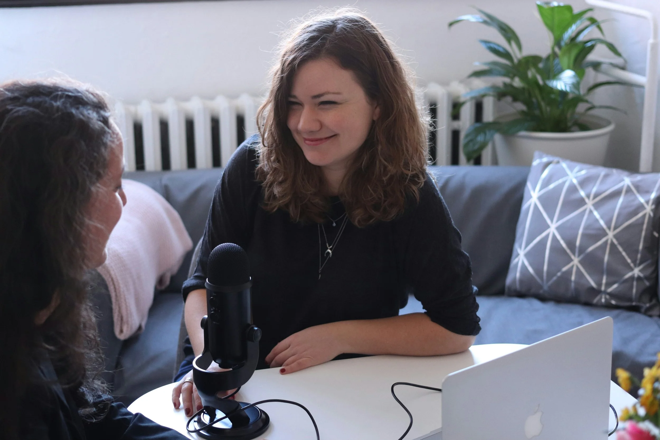 Two women engaging in a podcast or conversation, one speaking into a microphone, in a cozy room with a laptop, a sofa, pillow, and a potted plant in the background.