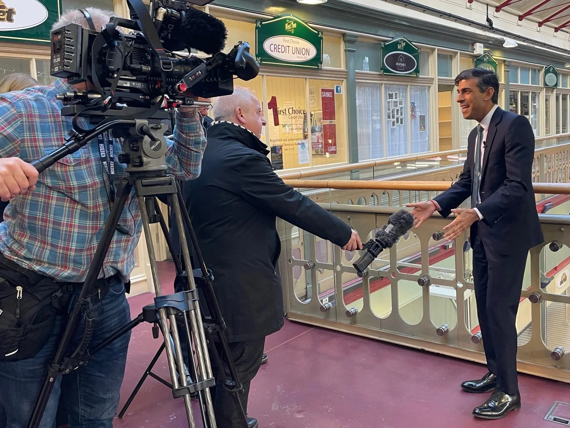 A news crew conducts an interview with a man in a suit at a shopping arcade. The man is smiling and gesturing with his hands. The crew includes a cameraman operating a large video camera and a person holding a microphone towards the interviewee. The setting features a balcony railing overlooking the lower level of the arcade, with storefronts and signs visible in the background.