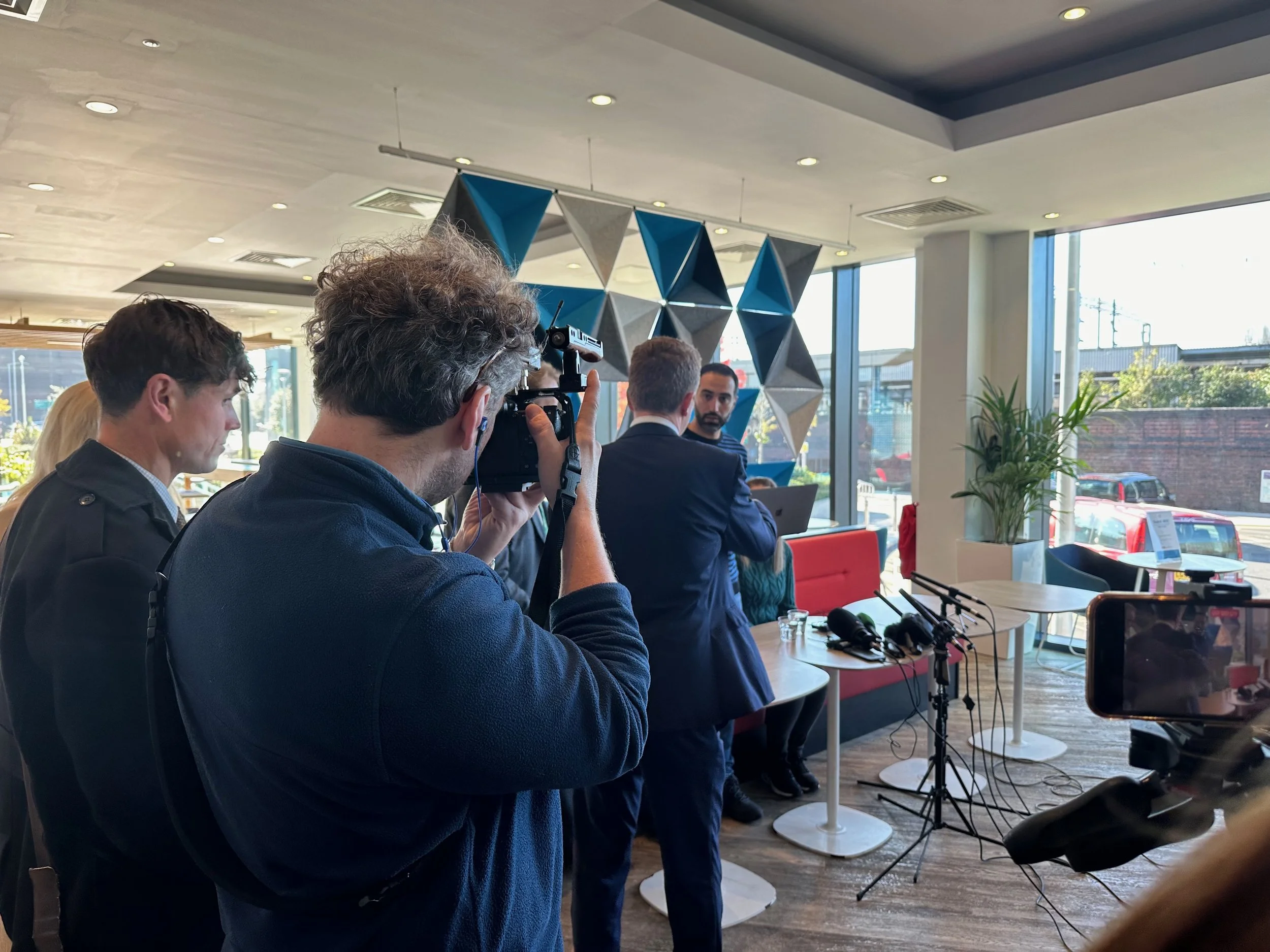 Man with curly hair taking a photo with a camera at a press event, with others and microphones on a table inside a room with large windows and decorative wall accents.