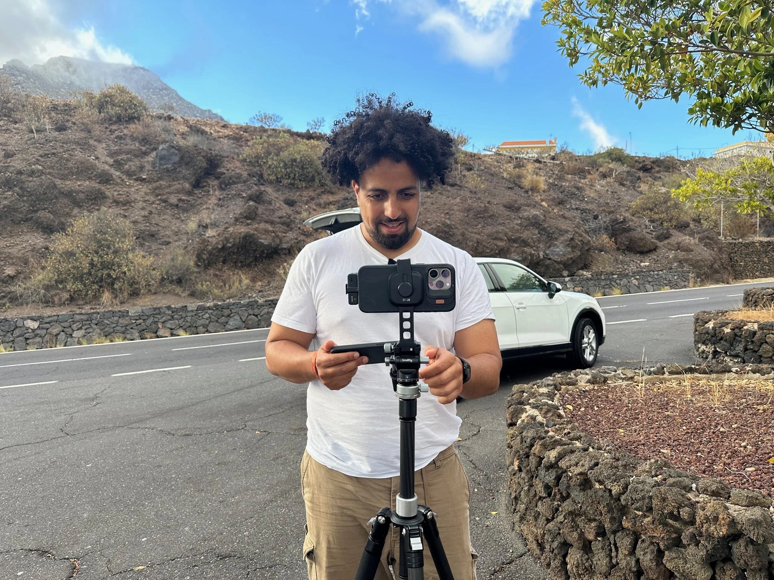 Man with curly hair operating a camera on a stabilizer outdoors with a rocky hillside, white car, and blue sky in the background.
