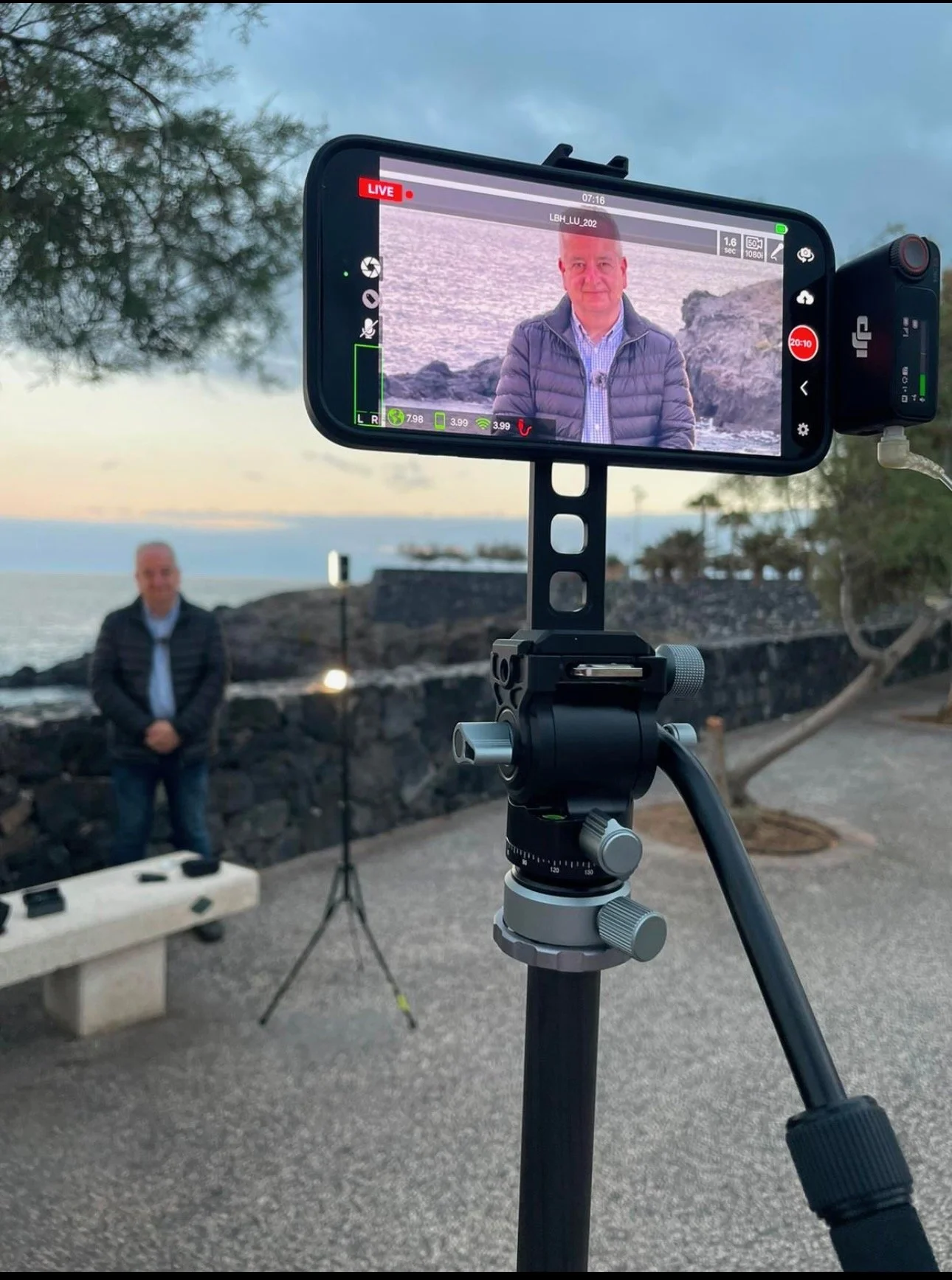 A person in a puffy jacket is being filmed by a smartphone mounted on a tripod near a waterfront during sunset. The screen of the smartphone shows the person facing the camera, with the ocean and trees in the background.