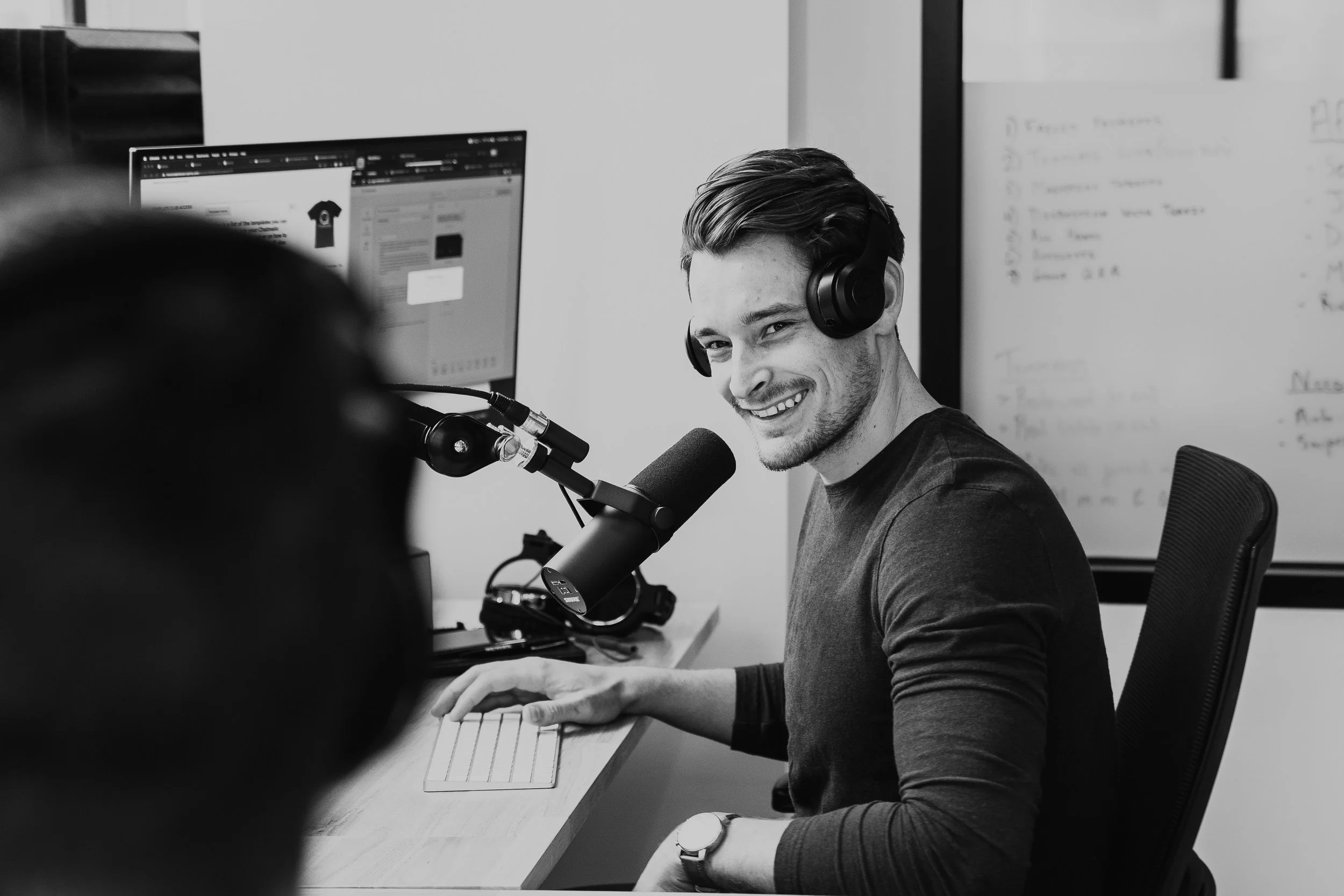 A young man with headphones smiling while speaking into a microphone in a podcast or radio studio, with a computer monitor and whiteboard in the background.