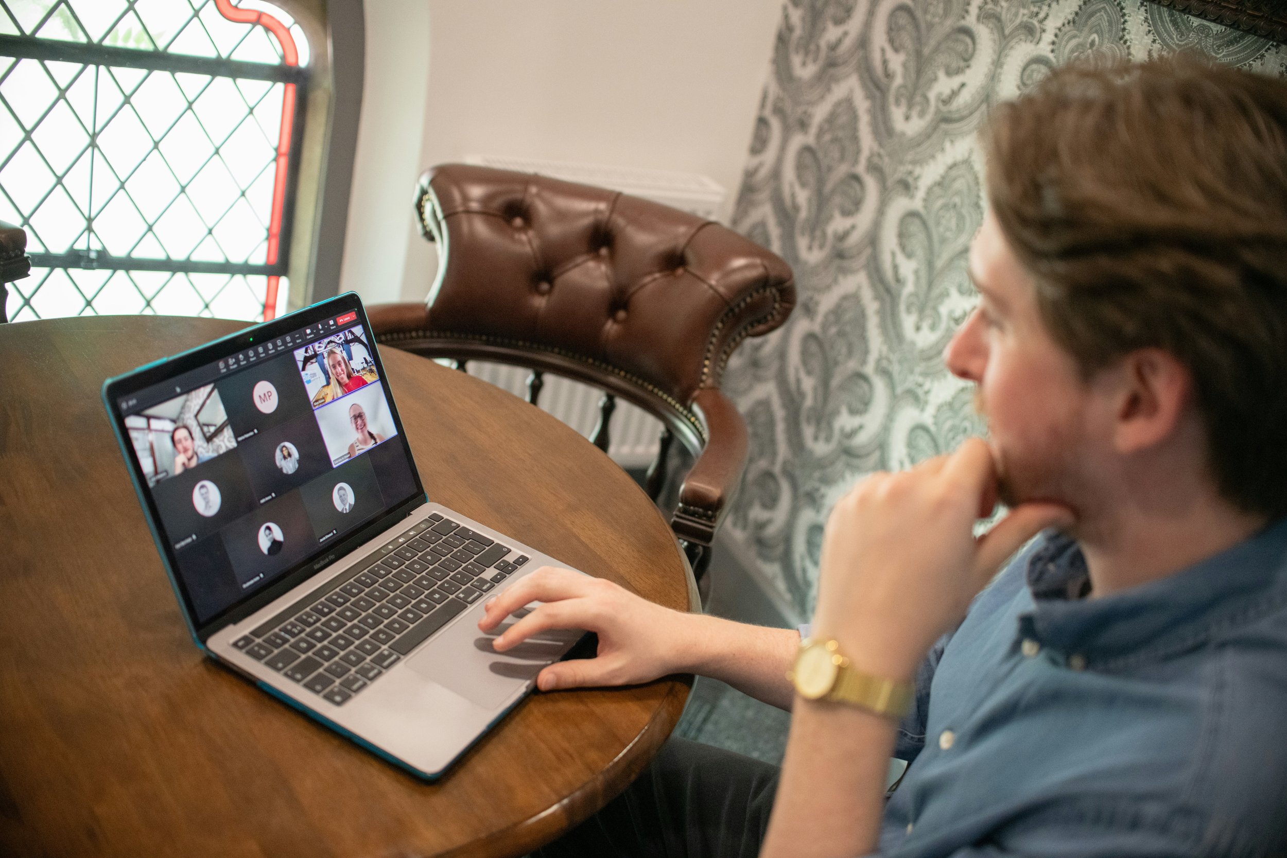 A man participating in a video conference on a laptop at a round wooden table in a room with patterned wallpaper, a leather armchair, and a stained glass window in the background.