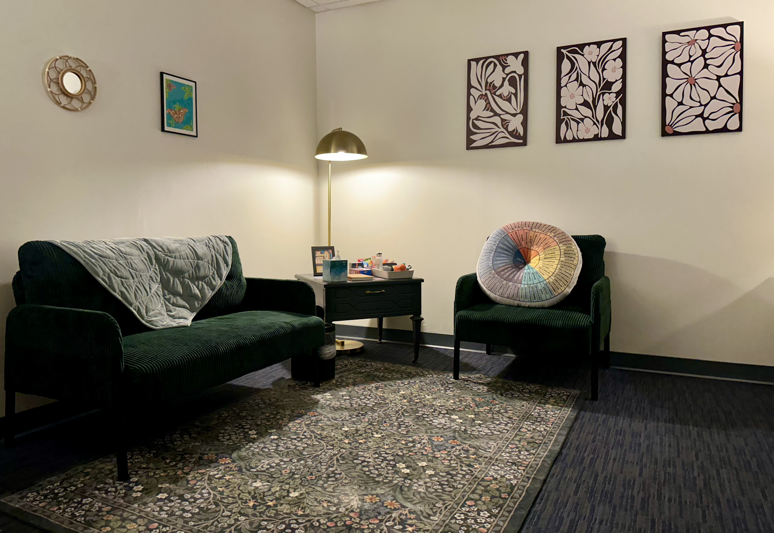 Picture of a counseling office with one green sofa and one green chair, a floral green rug, a gold lamp turned on, and various objects on a gray table (tissues, a framed picture, notebooks, and fidgets).