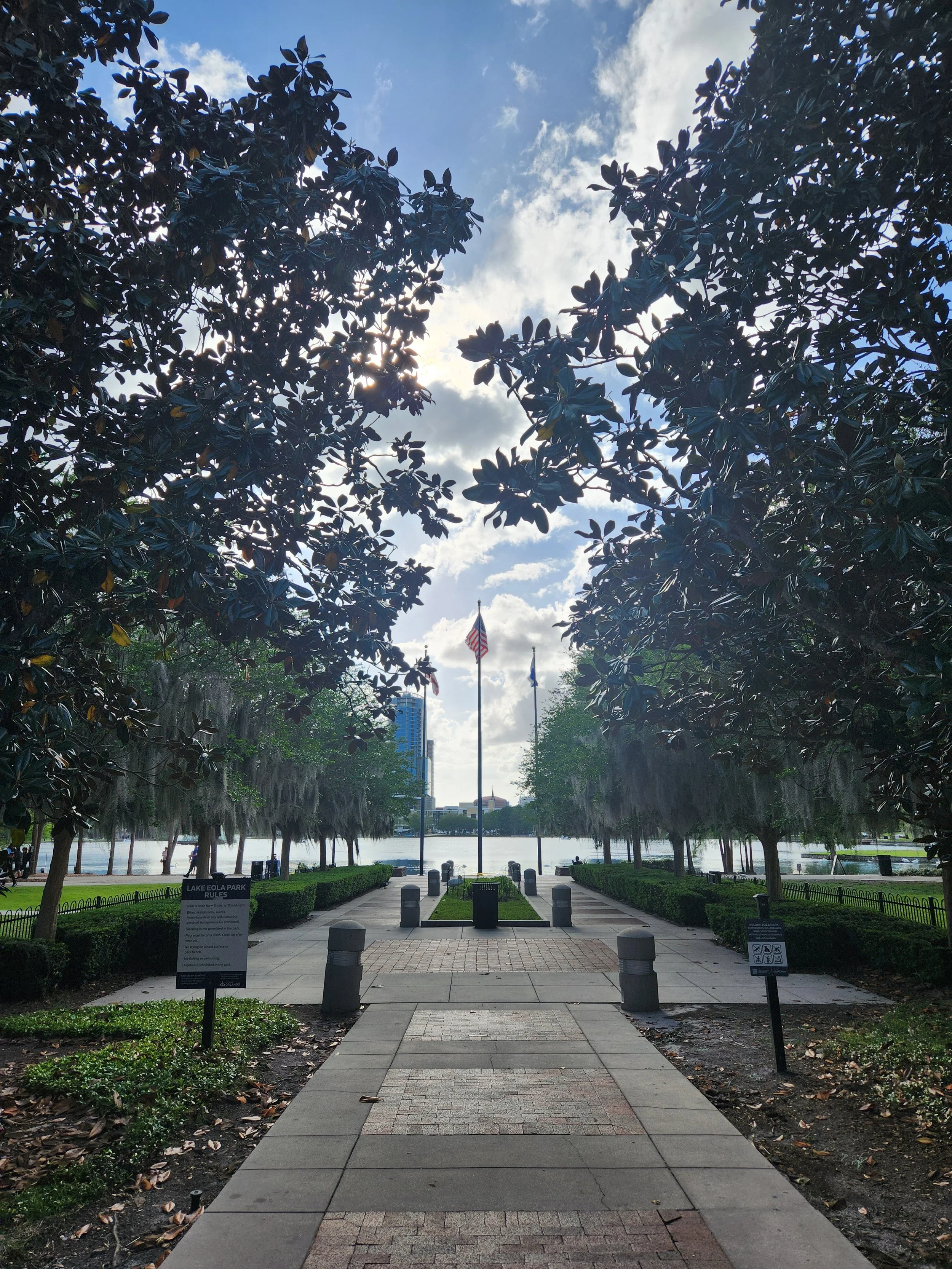 Pathway leading to a lake surrounded by trees in a park, with American flags flying on flagpoles, a city skyline visible in the background, and a partly cloudy sky overhead.