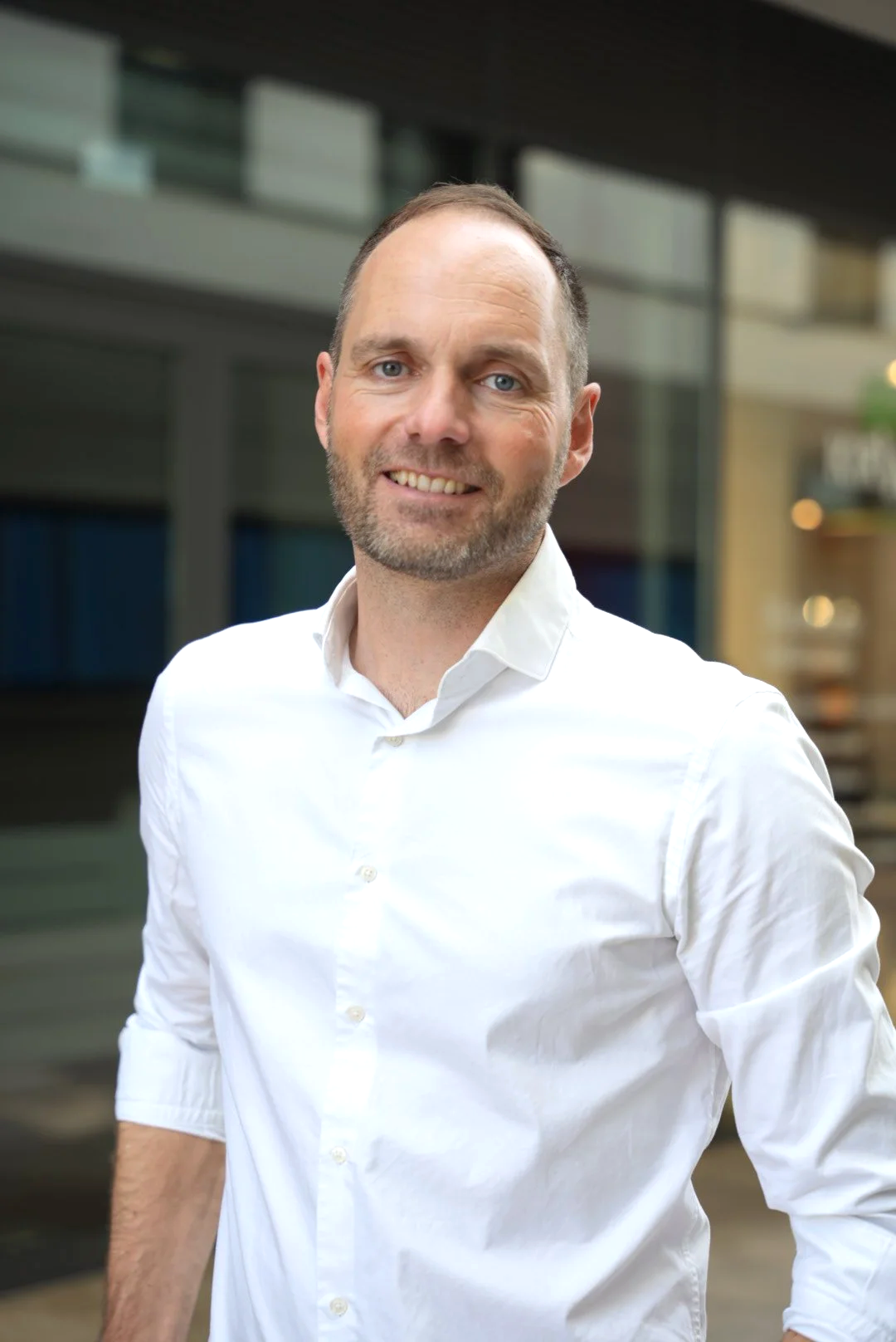 Porträt eines lächelnden Mannes in weißem Hemd vor einem modernen Gebäude in einer städtischen Umgebung.