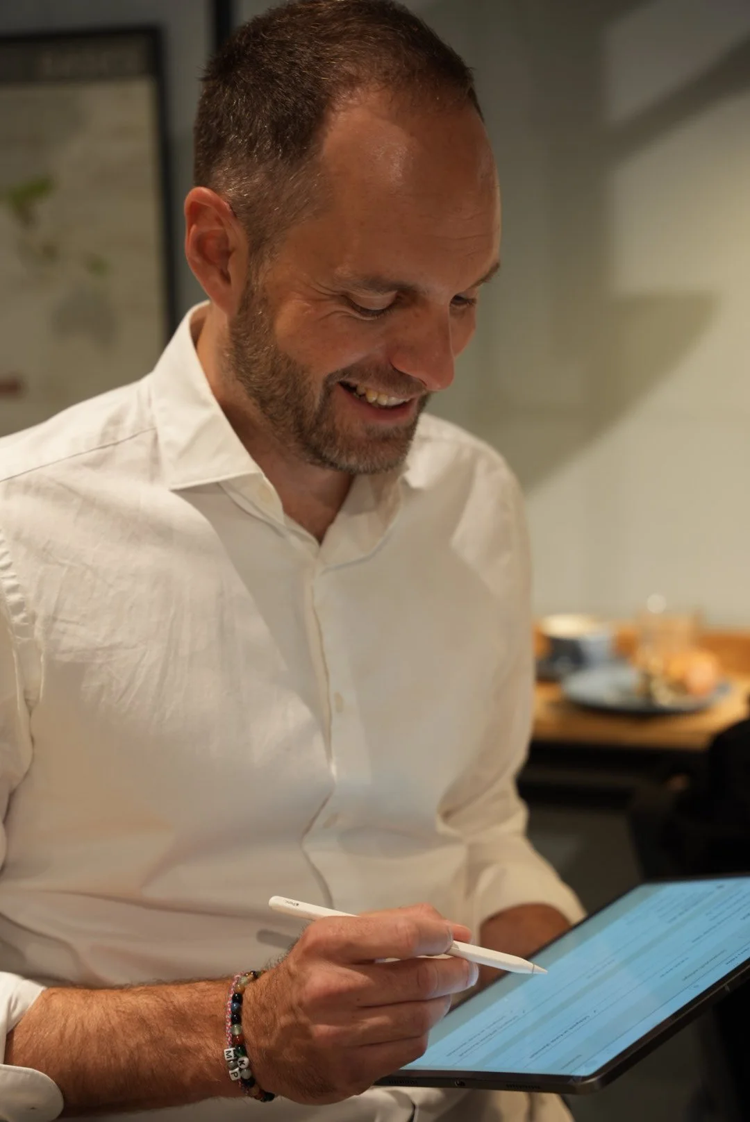 Ein Mann mit Bart und weißen Hemd, der auf einem Tablet mit einem Stylus schreibt oder zeichnet, lächelnd in einem Raum.