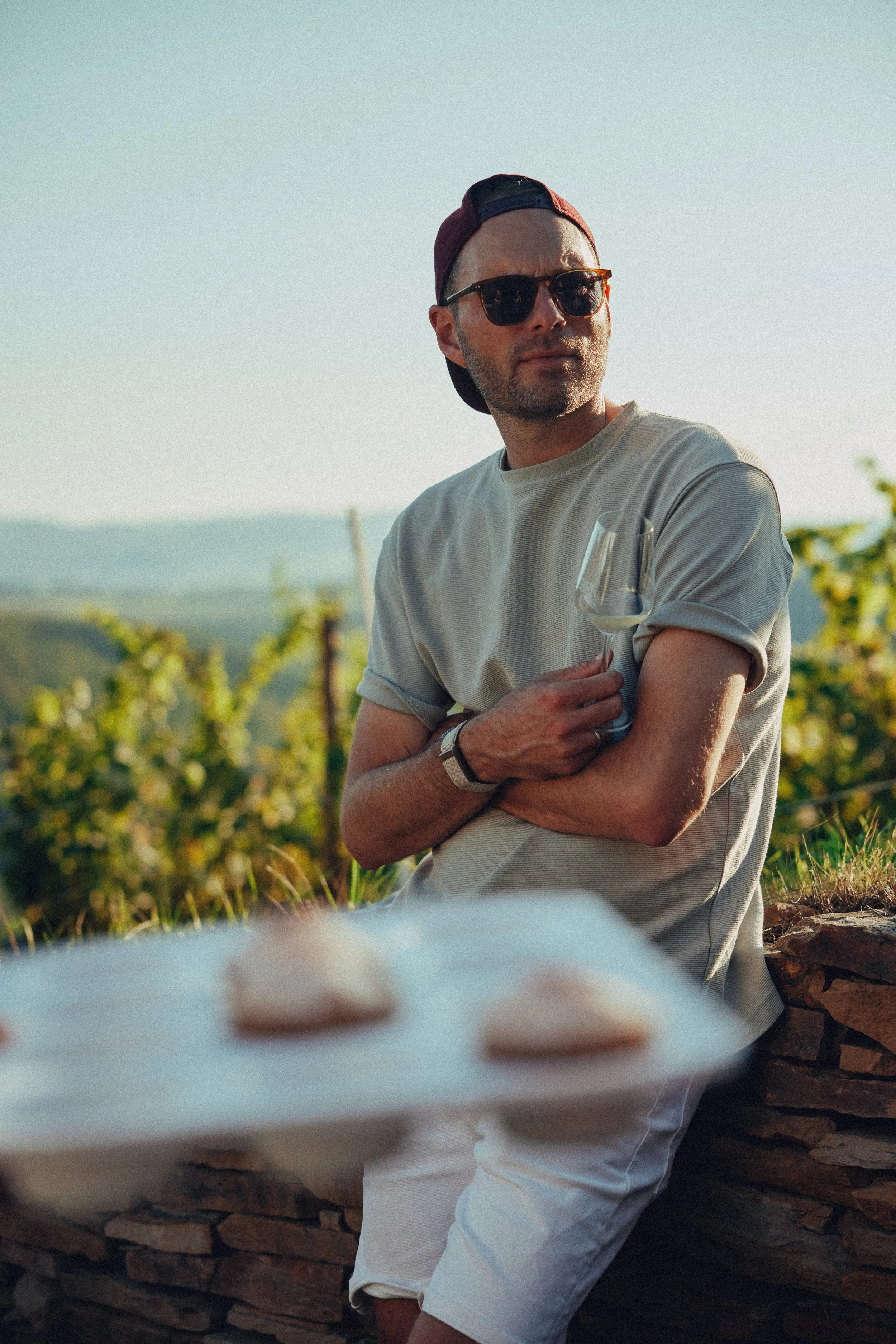 Sven-Ulrik Schneider mit Sonnenbrille, Baseballcap und einem T-Shirt steht bei Sonnenuntergang in einer Weinberg-Landschaft und hält ein Weinglas in der Hand, während im Vordergrund eine Platte mit Keksen leicht unscharf zu sehen ist.