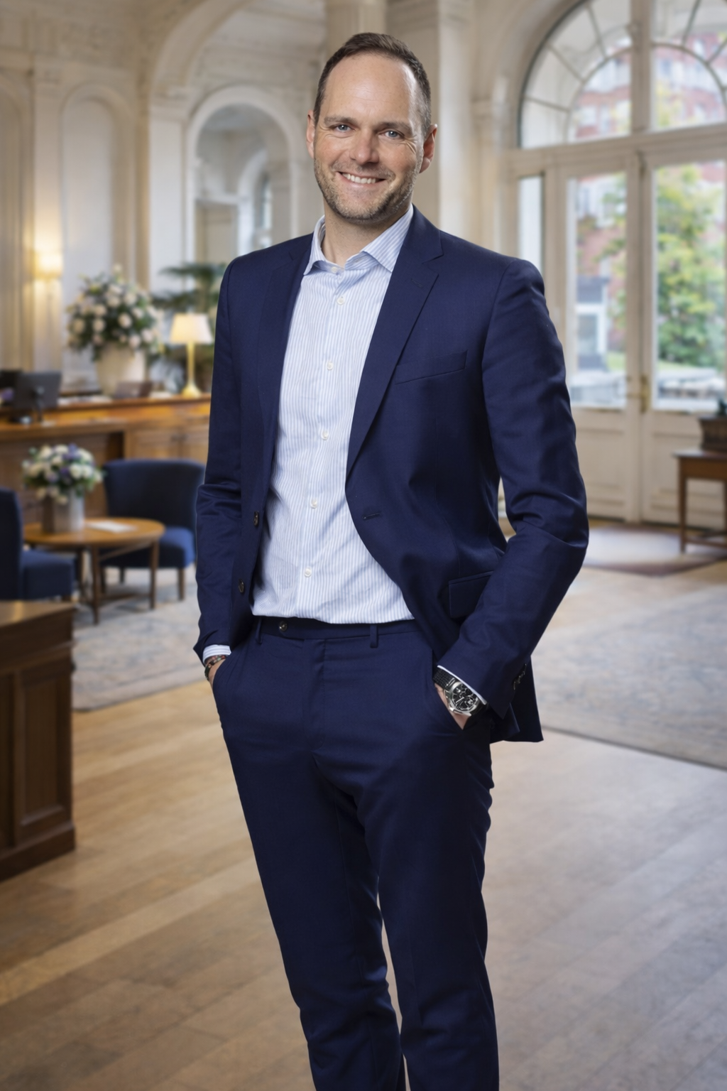 Ein Mann in einem dunkelblauen Anzug und hellblauem Hemd steht lächelnd in einem eleganten Raum mit großen Fenstern, Holzinterior und Blumen im Hintergrund.