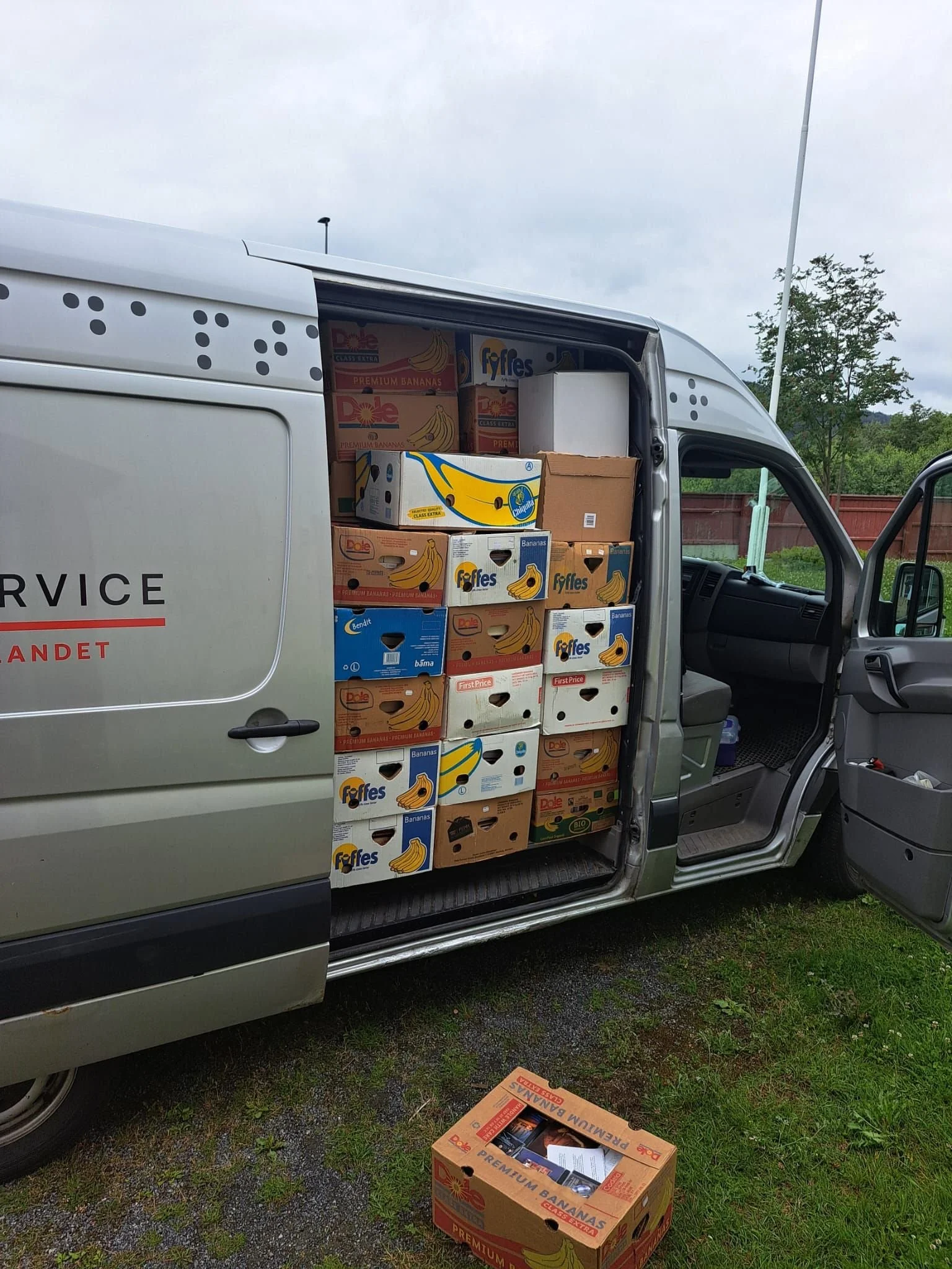 Varebil med esker med bananer, plassert i lasteområdet, med en åpen bakdør og en åpen passasjerdør, plassert på gress. En ekstra eske ligger på bakken utenfor bilen.
