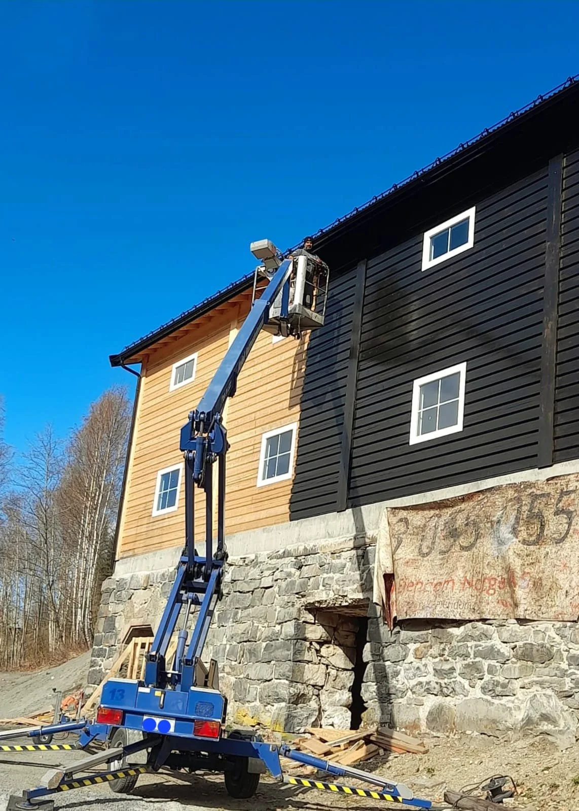 En person står i en lift foran en bygning med tre etasjer, hvor den ene delen er kledd med trepanel og den andre med svart panel. Bygningen står på en grunnmur av store steinblokker.