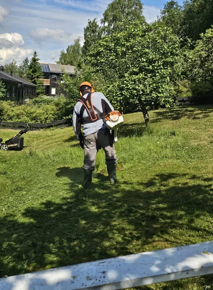 Person iført hjelm, vernebriller, strykejern og støvler som bruker gressklipper i hagen.