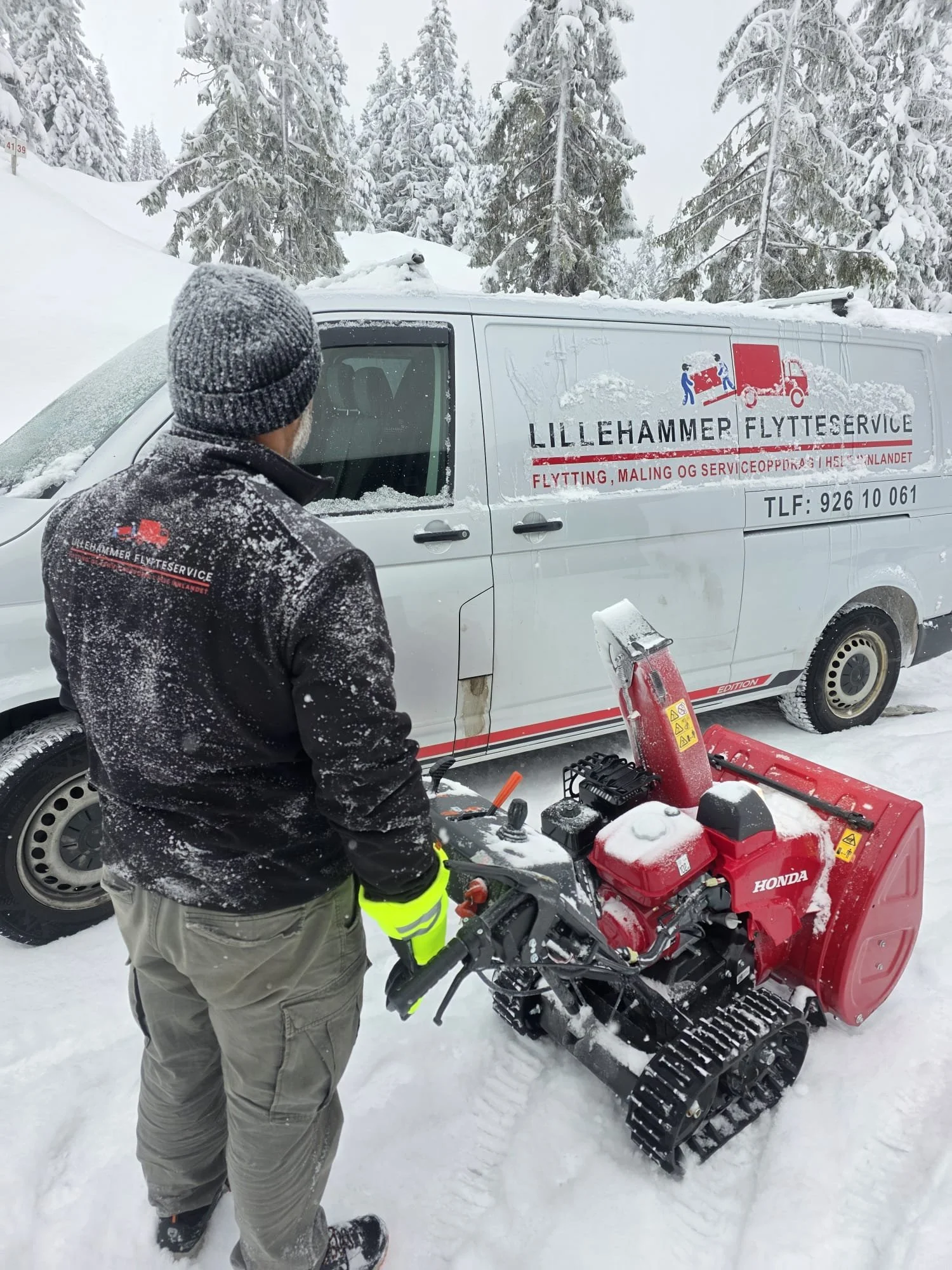 En person står i snø med en høytrykksspyler foran en hvit varebil fra Lillehammer Flytteservice, omgitt av snødekt landskap og trær.
