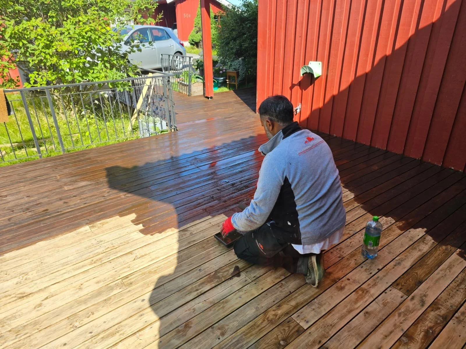 En mann som legger nytrete treplanker på en terrasse utendørs, mens solen skinner og det er grønt landskap og en parkert bil i bakgrunnen.