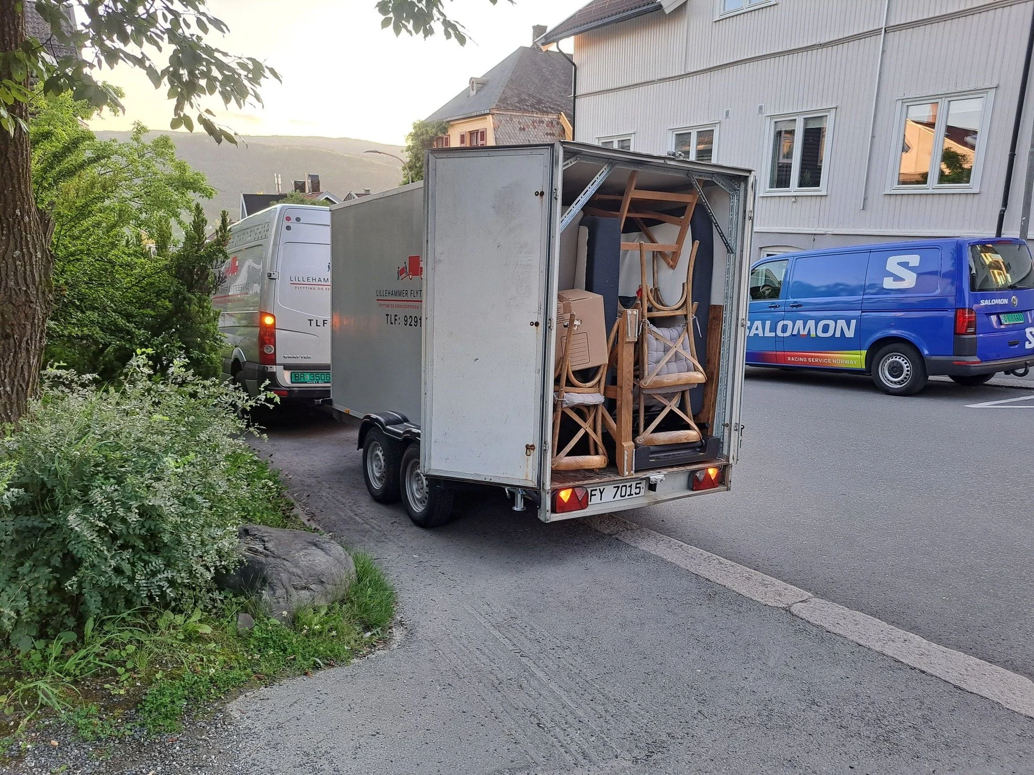 En liten trailer fylt med stoler og småmøbler, parkert på gaten ved siden av andre kjøretøy i en liten bygd.