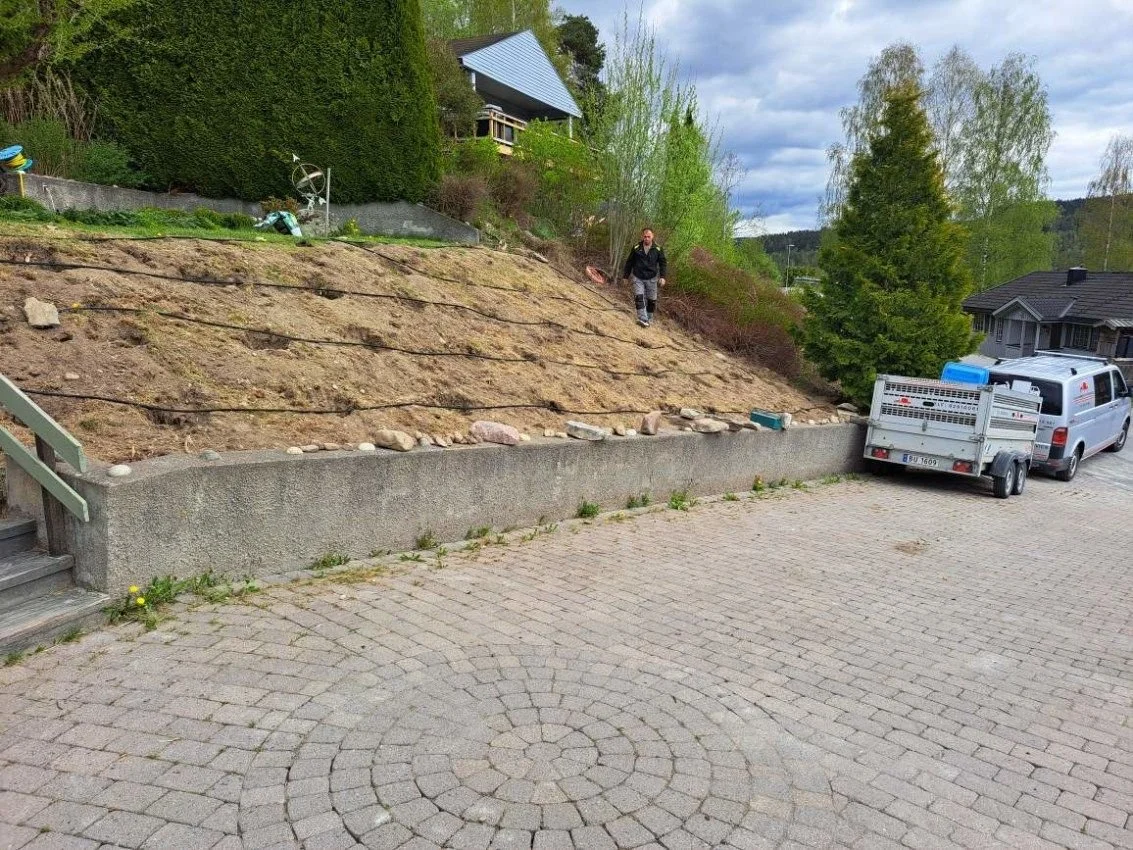 En person står på en skråning med jord og steiner, som er under arbeid, ved en vei med brostein. Det er trær og hus i bakgrunnen.