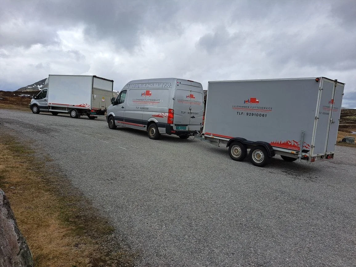 Tre flyttebiler parkert på en grusvei i et vakkert, åpent landskap med fjell i bakgrunnen og overskyet vær.