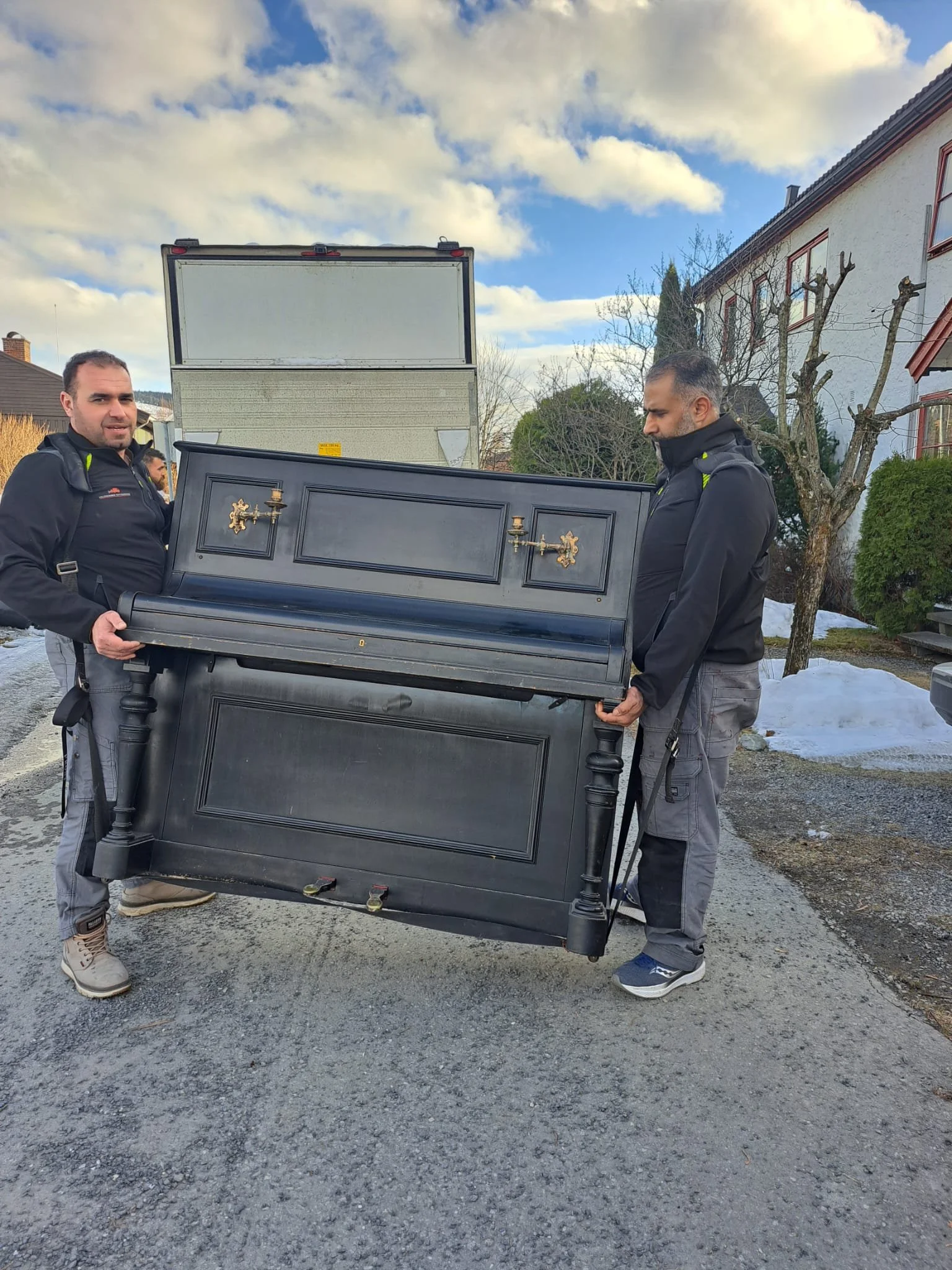 To menn bærer en svart og mørk grå gammel piano på en gate med hus og trær i bakgrunnen.