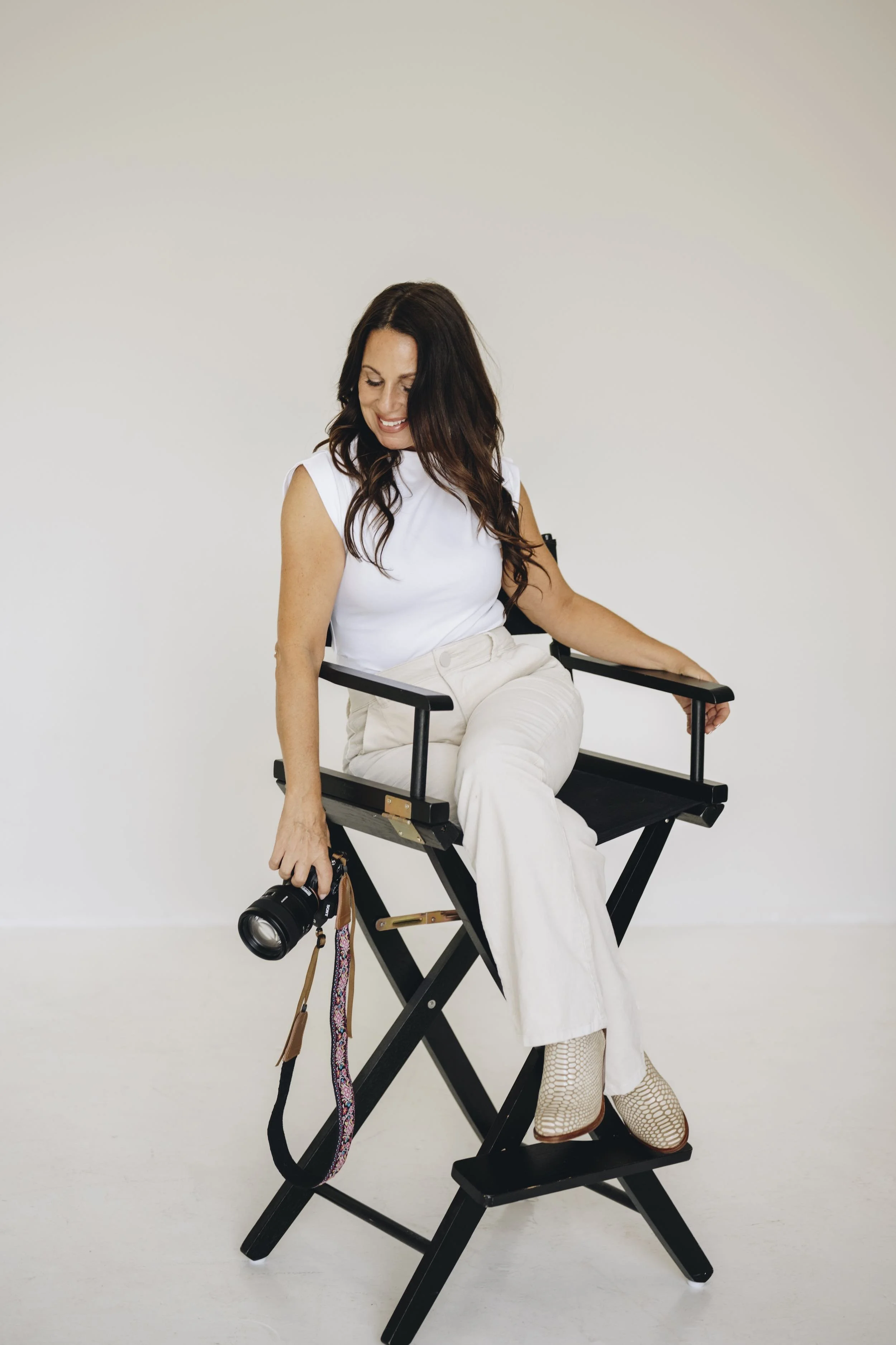 A woman with dark brown hair wearing a white top and cream-colored pants sitting on a black director's chair against a plain white wall. She is smiling, looking down, and holding a professional camera with a strap hanging down.