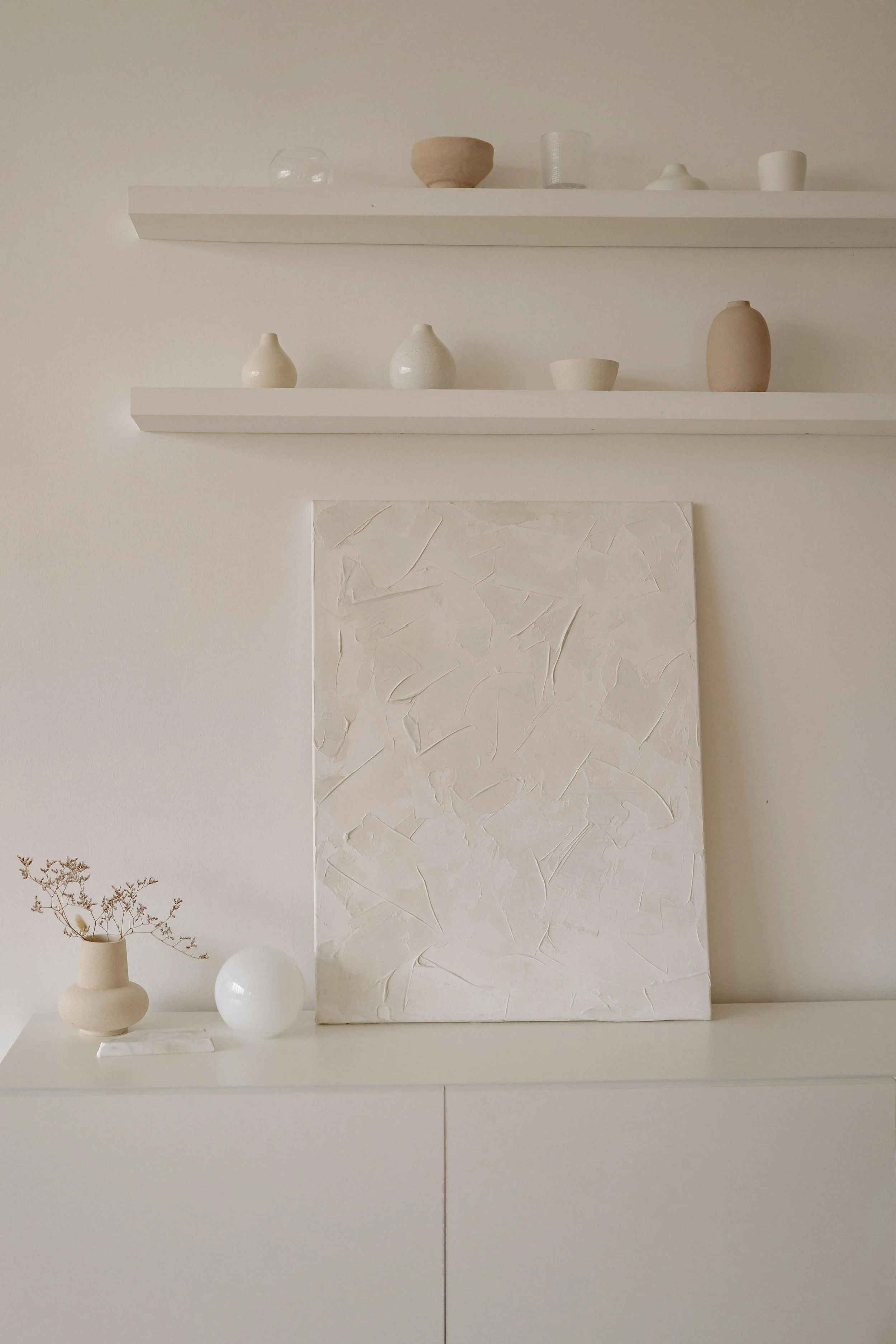 Minimalist interior with two white shelves holding small neutral-colored vases and bowls, a textured white canvas on the wall, and a table with decorative items including a vase with dried flowers and two white spheres.