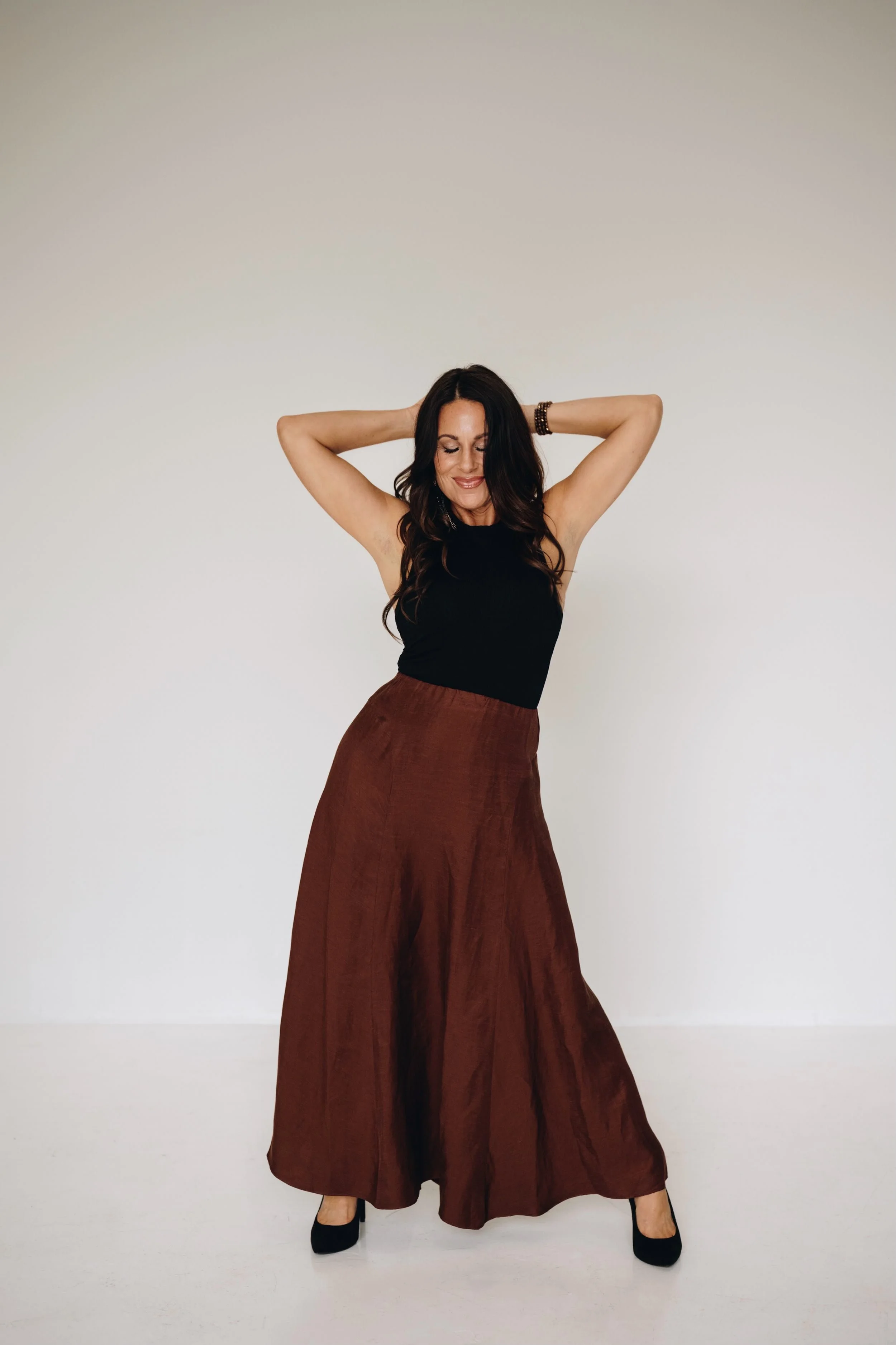 Woman with long dark hair in a black sleeveless top and brown wide-leg pants, standing with arms raised and eyes closed against a plain white background.