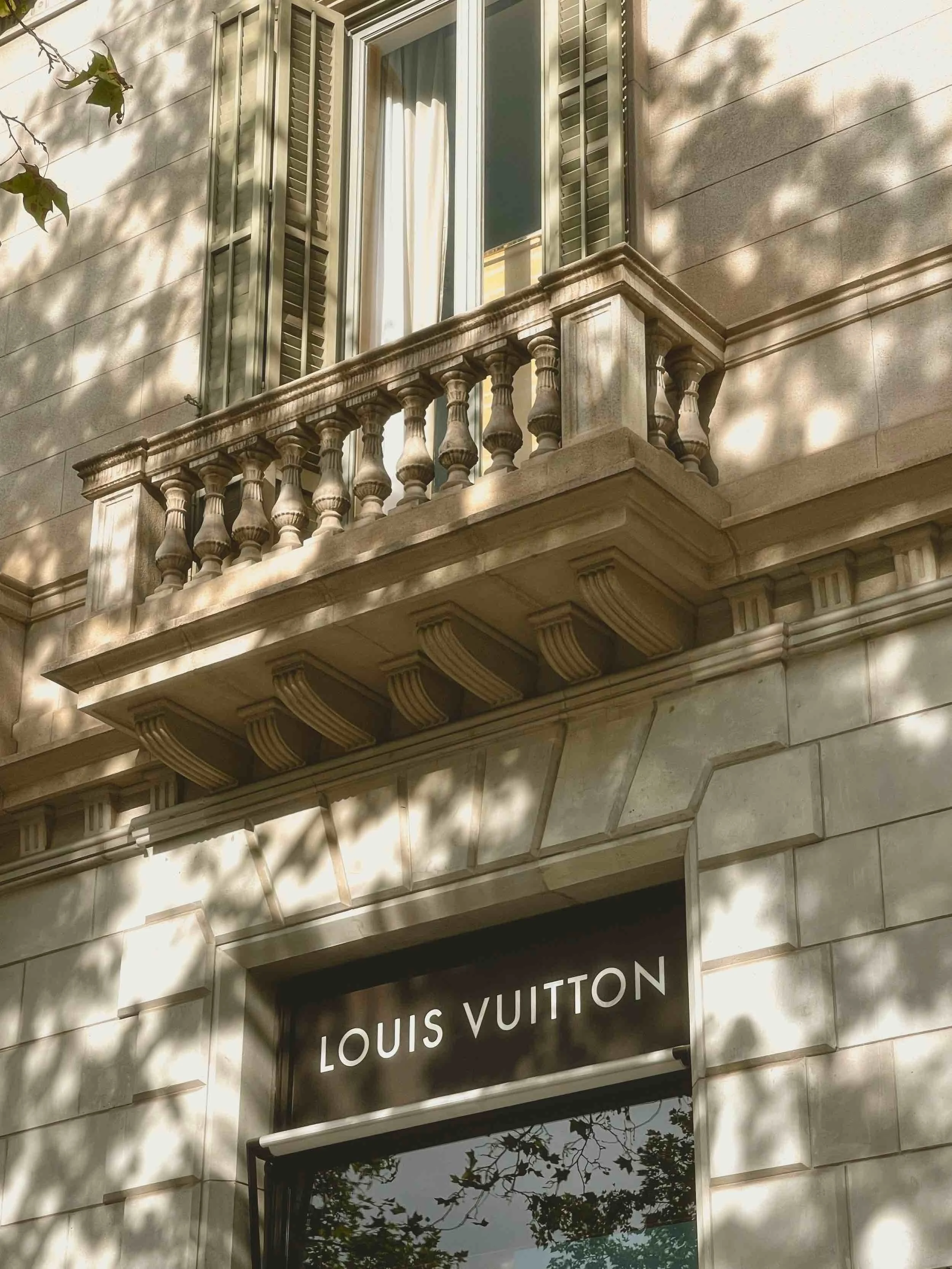 Exterior view of a Louis Vuitton store with a small balcony with stone balustrades and a window with open shutters. The store's sign is visible below the balcony, with the brand name clearly displayed.