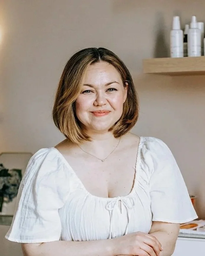 A woman with shoulder-length brown hair and a white puff-sleeve blouse smiling, standing indoors with shelves and bottles in the background.