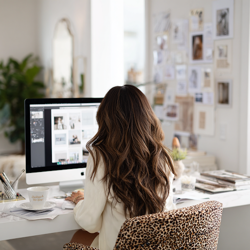 A woman with long wavy brown hair working on a computer in a bright, decorated home office with photos on the wall.