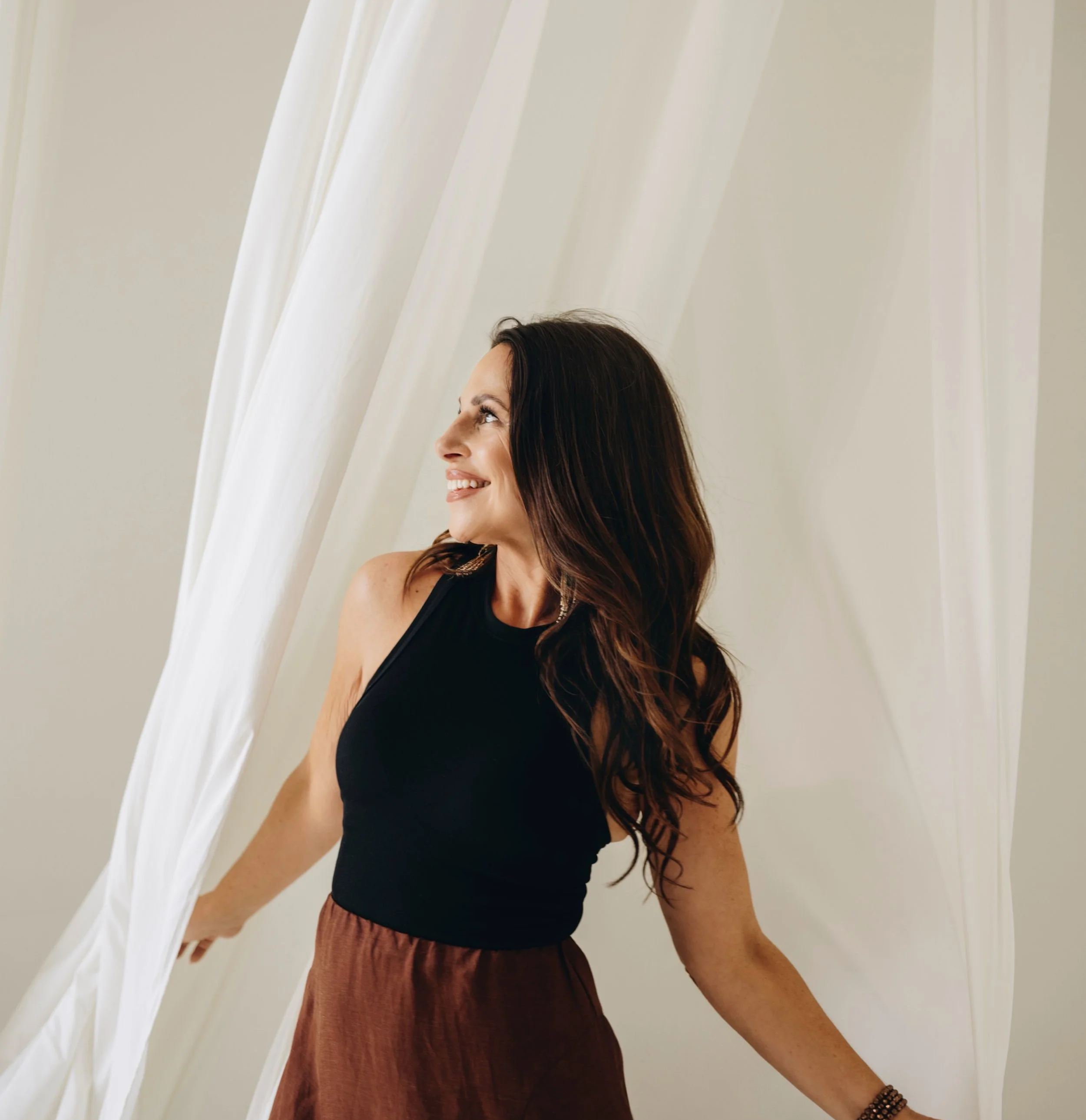 Profile of a smiling woman with long dark hair looking upward inside a room with white draped curtains.
